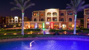 Een hotel met een zwembad, palmbomen en een verlichte ingang.