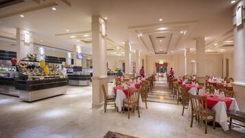 A hotel dining area with multiple tables set for guests, a buffet with various food items, and a television screen mounted on the wall.
