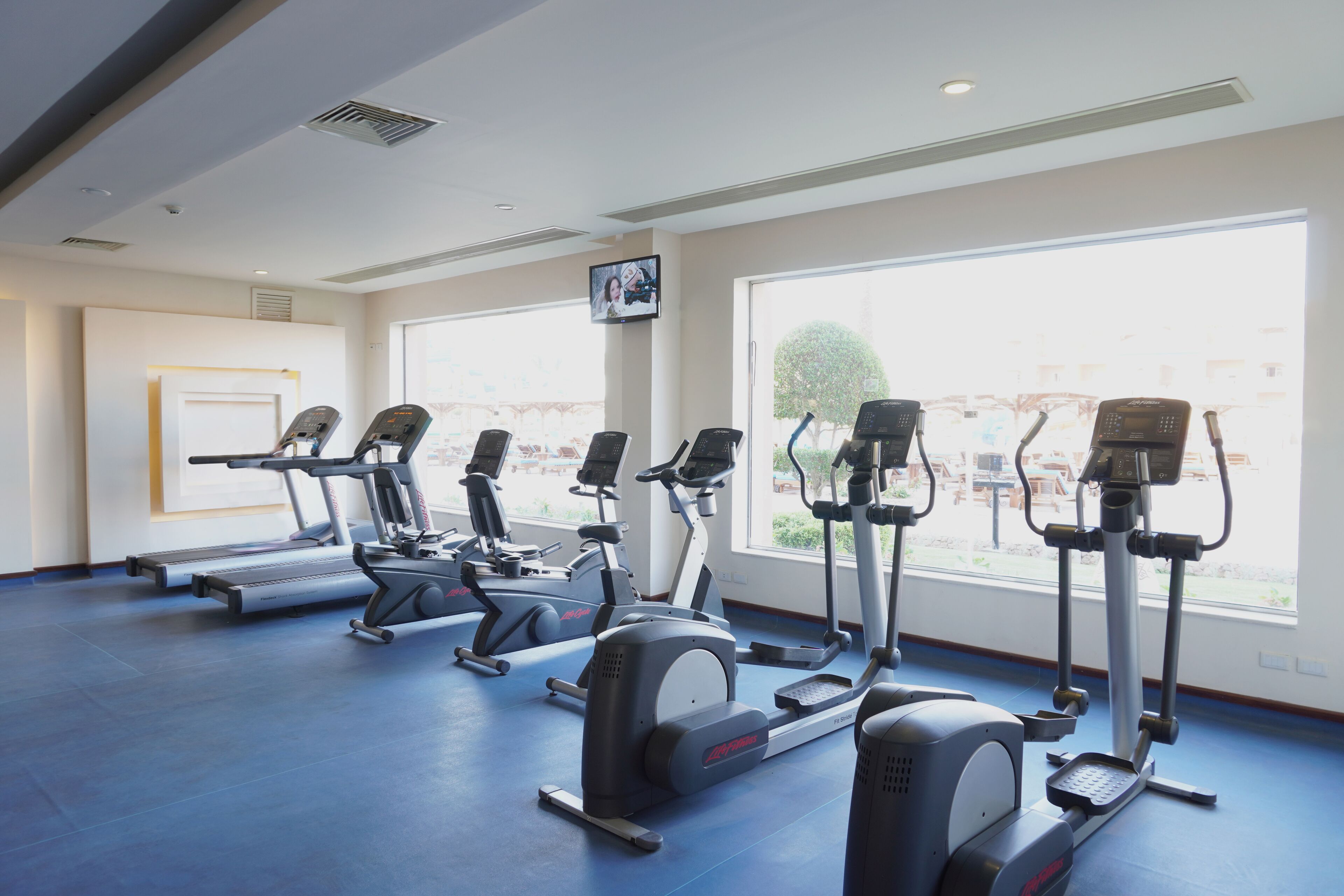 Un gimnasio con varias máquinas de cardio, ventanas amplias y vistas al exterior.