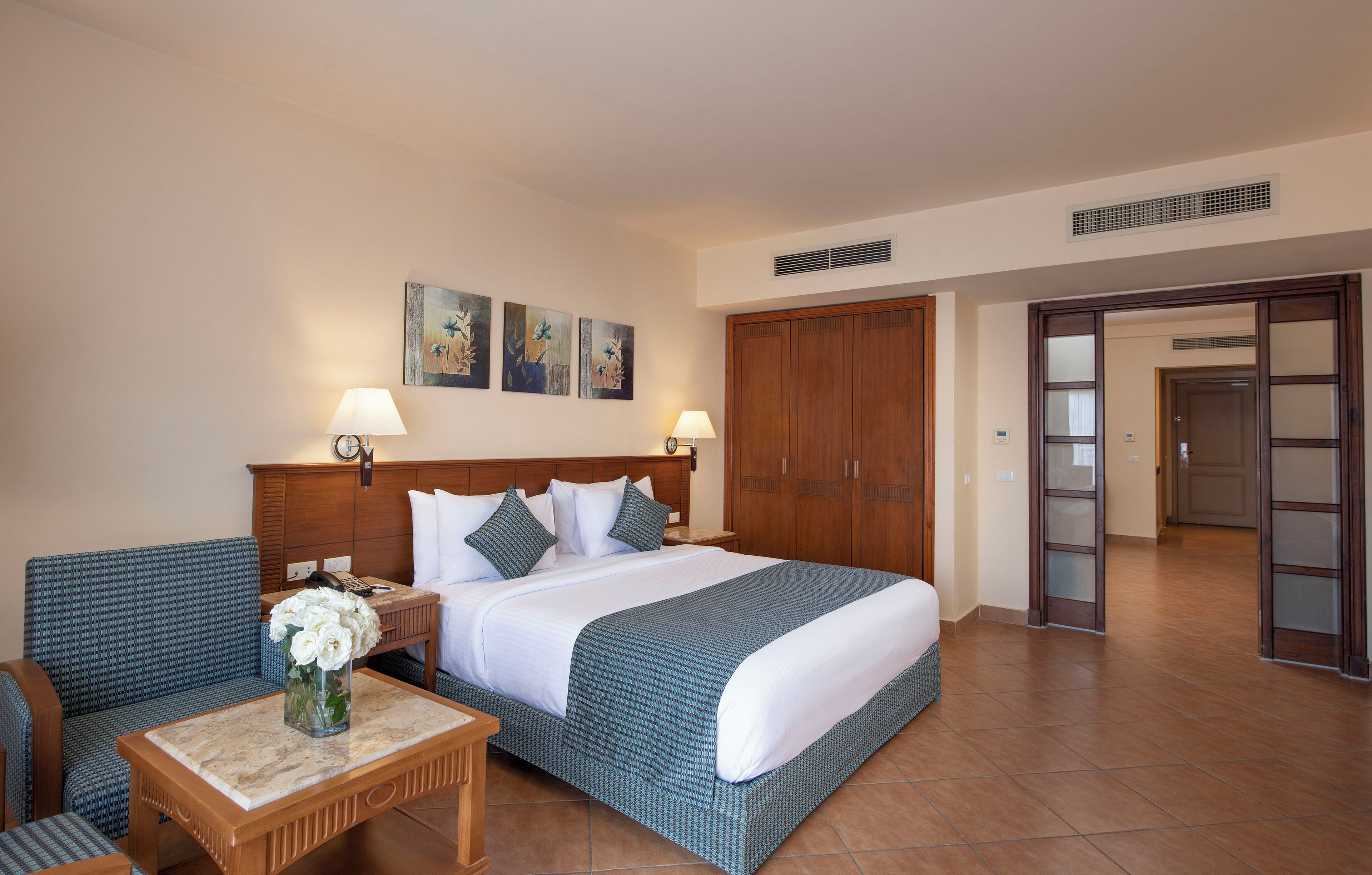 Une chambre d’hôtel avec un grand lit, une tête de lit en bois, deux tables de chevet avec des lampes, une petite table sur laquelle est posé un vase de fleurs et un fauteuil.