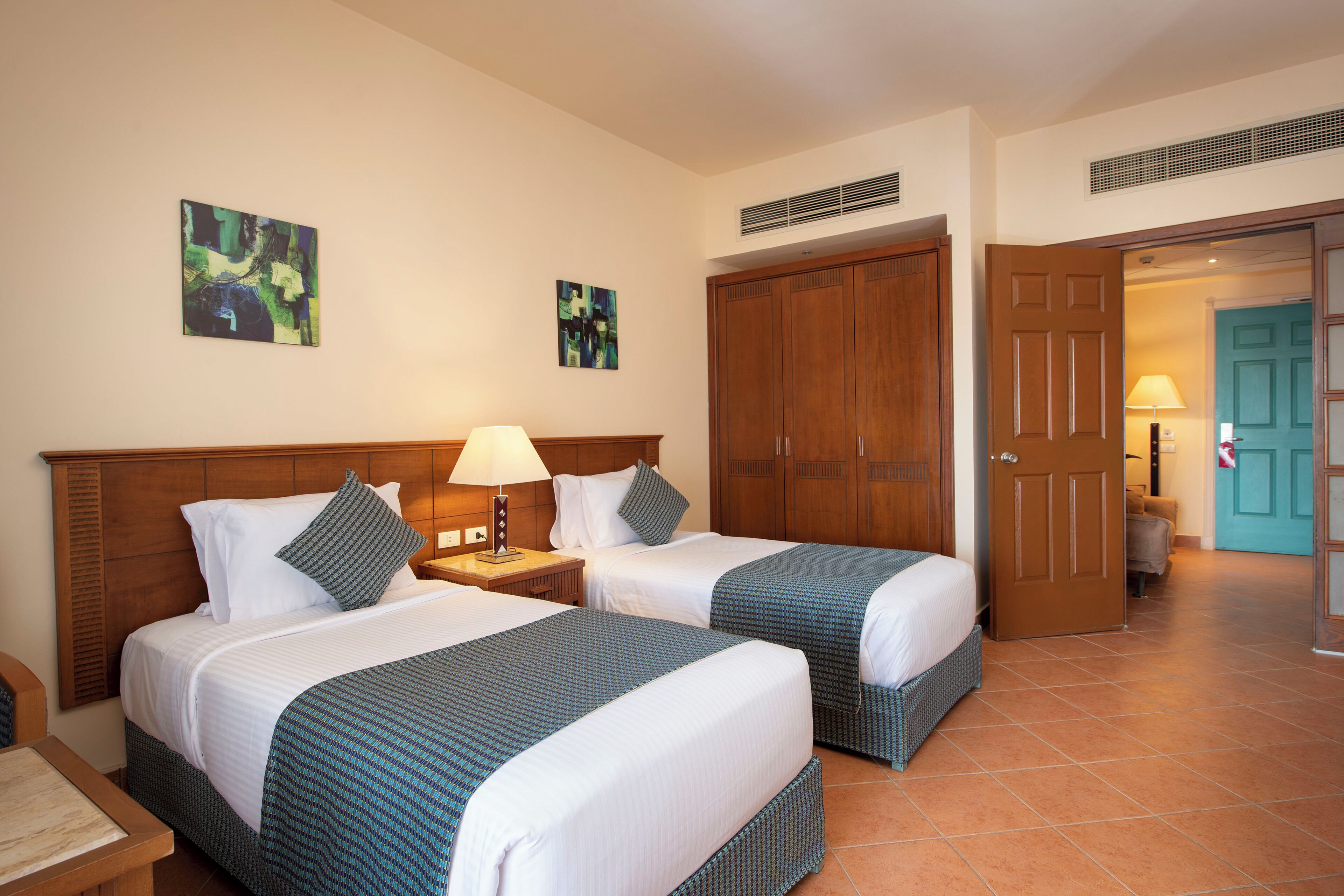 A hotel room with two beds, a wooden wardrobe, a door leading to another room, and artwork on the walls.
