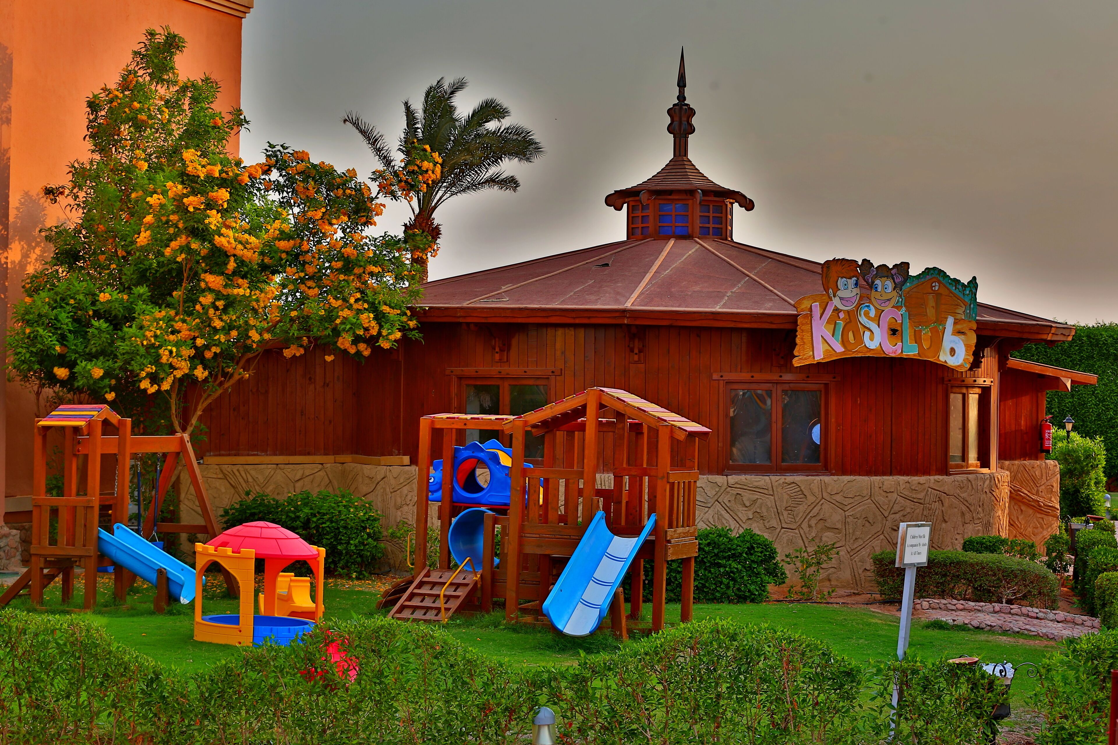 A wooden playhouse with a slide and climbing area, surrounded by greenery and a cloudy sky.