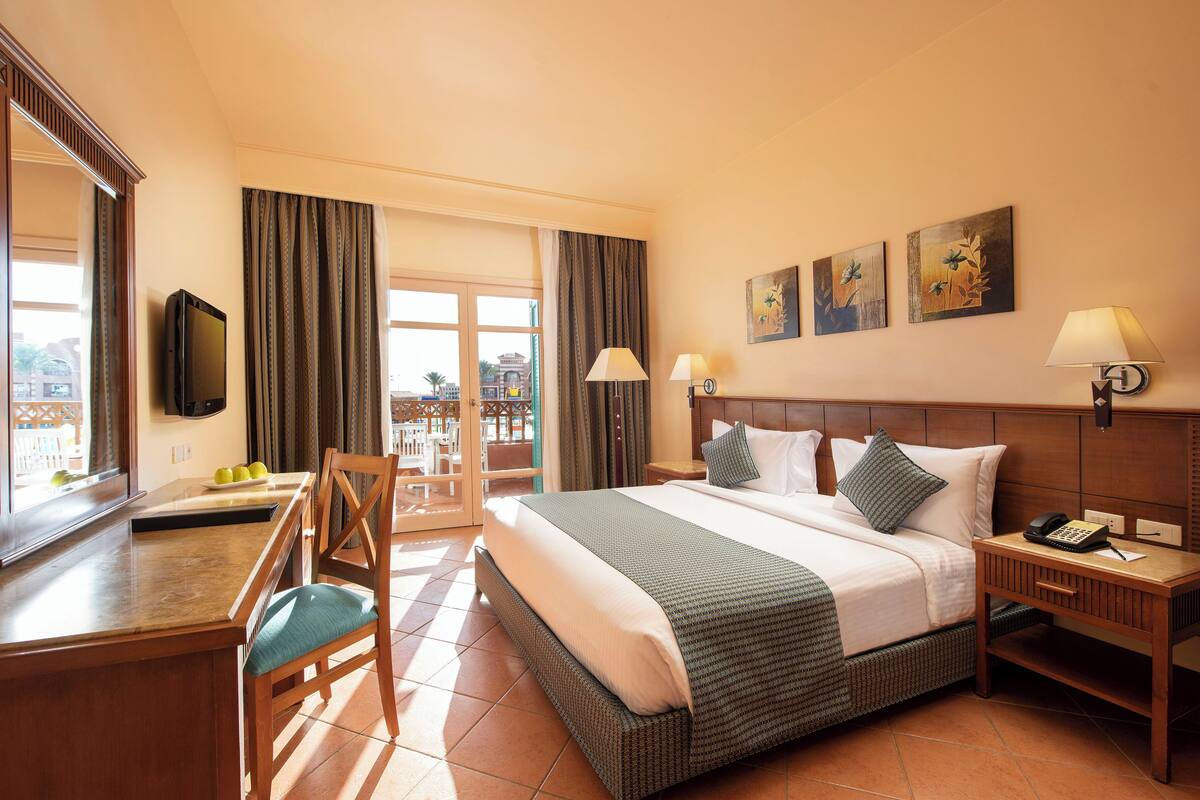 Une chambre d’hôtel avec un grand lit, un bureau avec une chaise, une télévision et un balcon avec des rideaux.