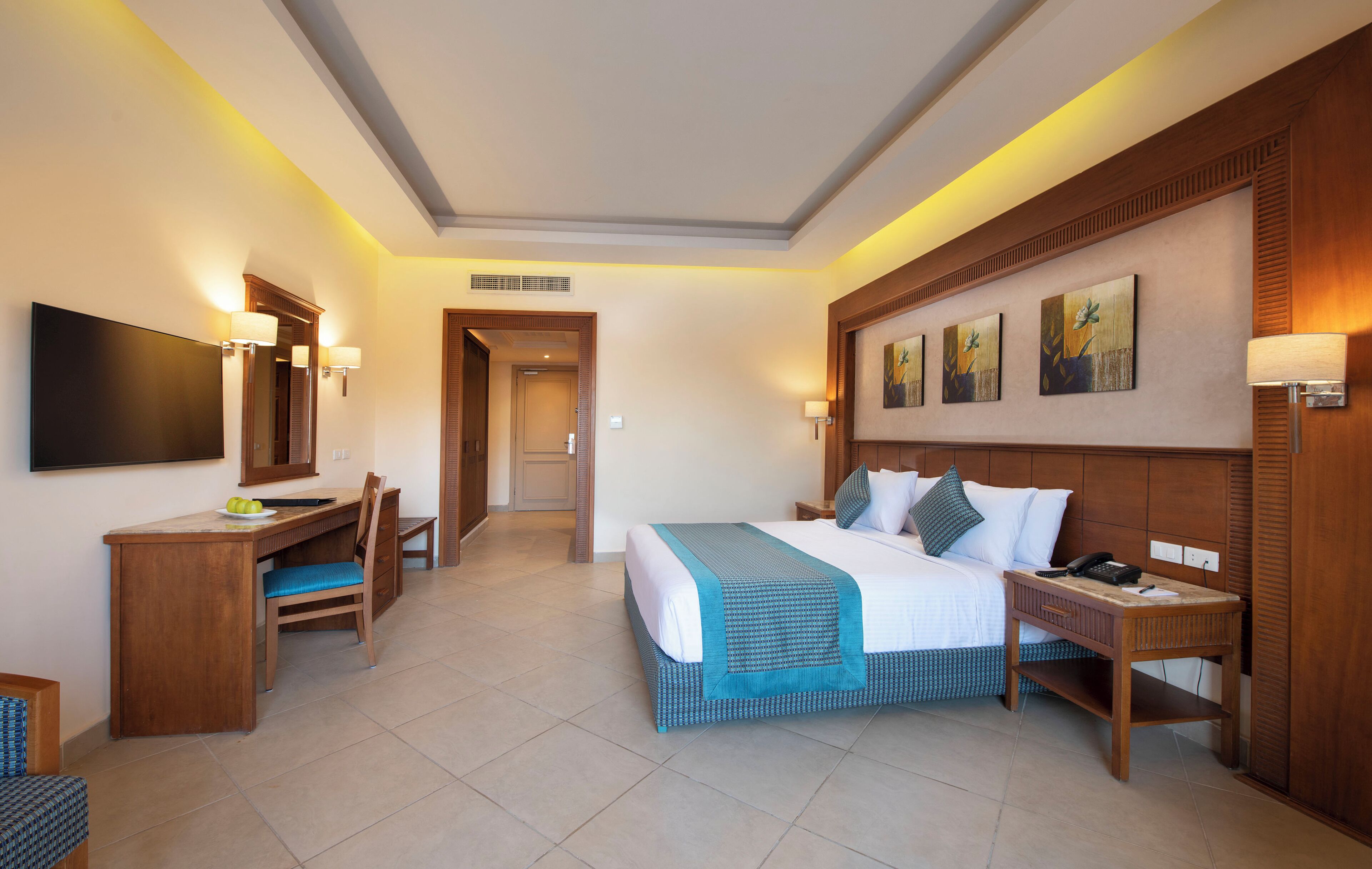 Ein Hotelzimmer mit Bett, Schreibtisch, Stuhl, Nachttisch, Lampe, Kunstwerk und Fernseher.