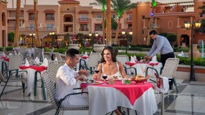 Een terras met tafels voor twee personen, omringd door palmbomen en een gebouw met balkons.