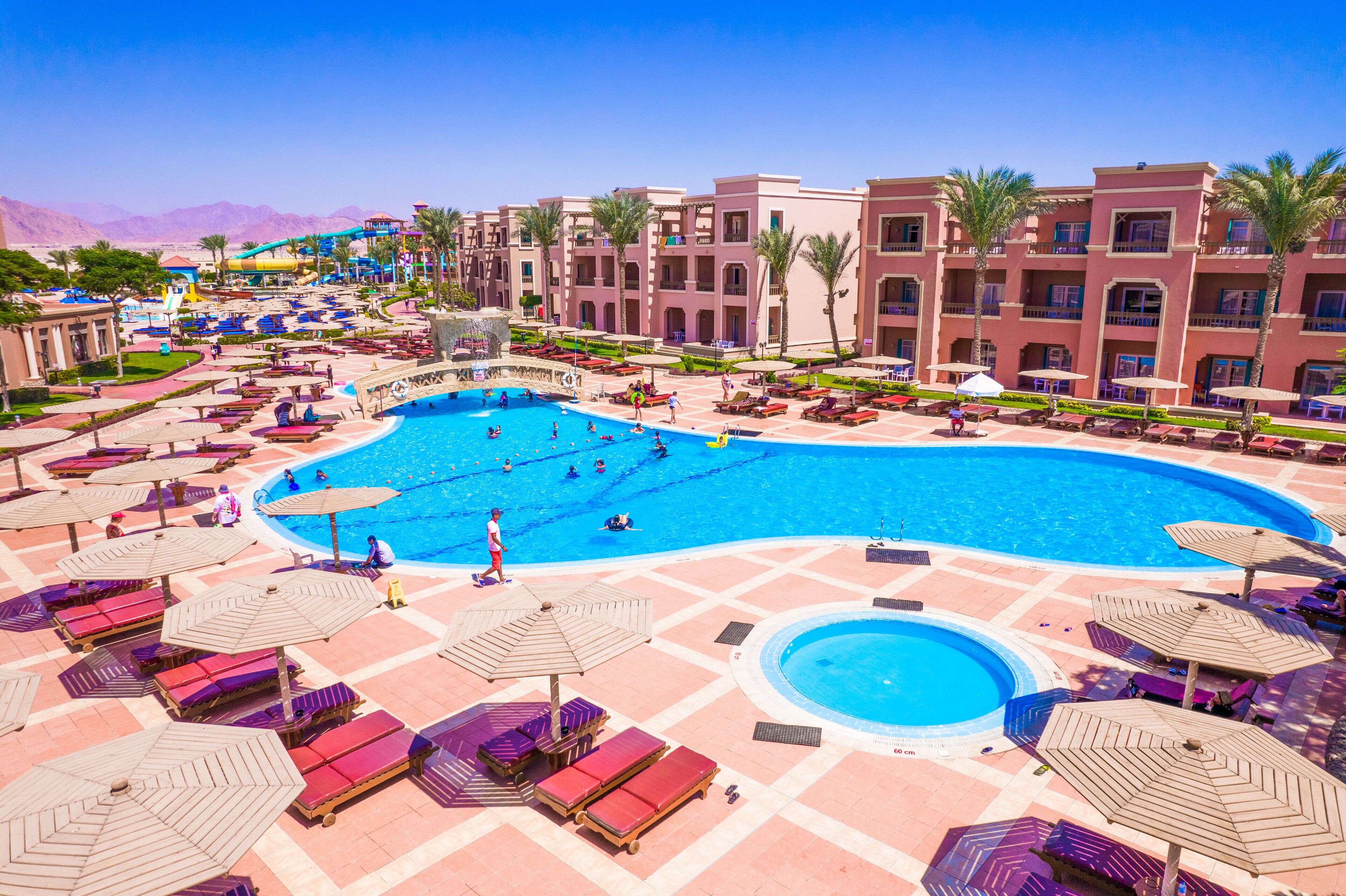 A large swimming pool with multiple lanes, surrounded by lounge chairs and umbrellas, and a water slide in the background.