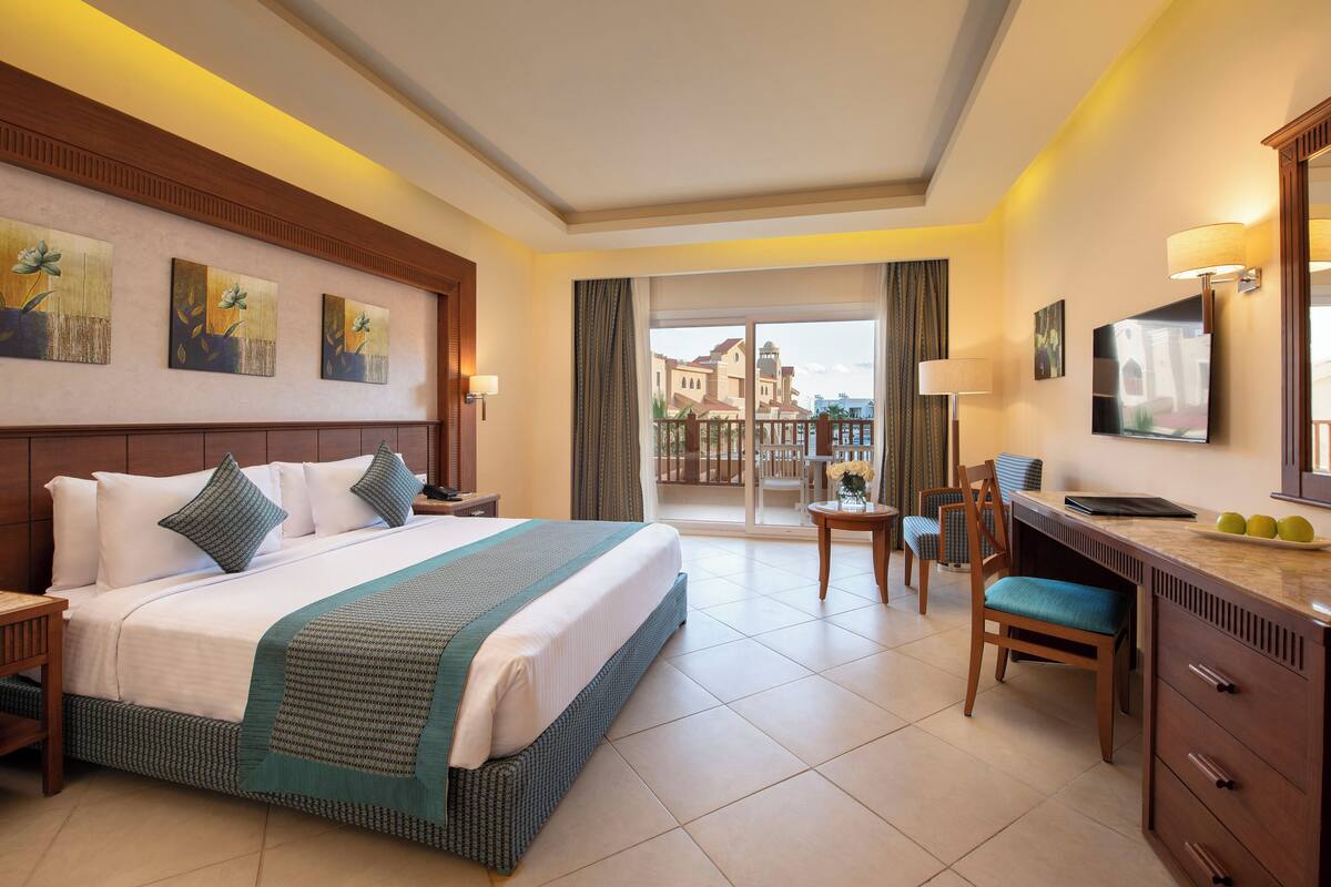 Une chambre d’hôtel avec un grand lit, un bureau, une chaise, une télévision et un balcon donnant sur des immeubles.