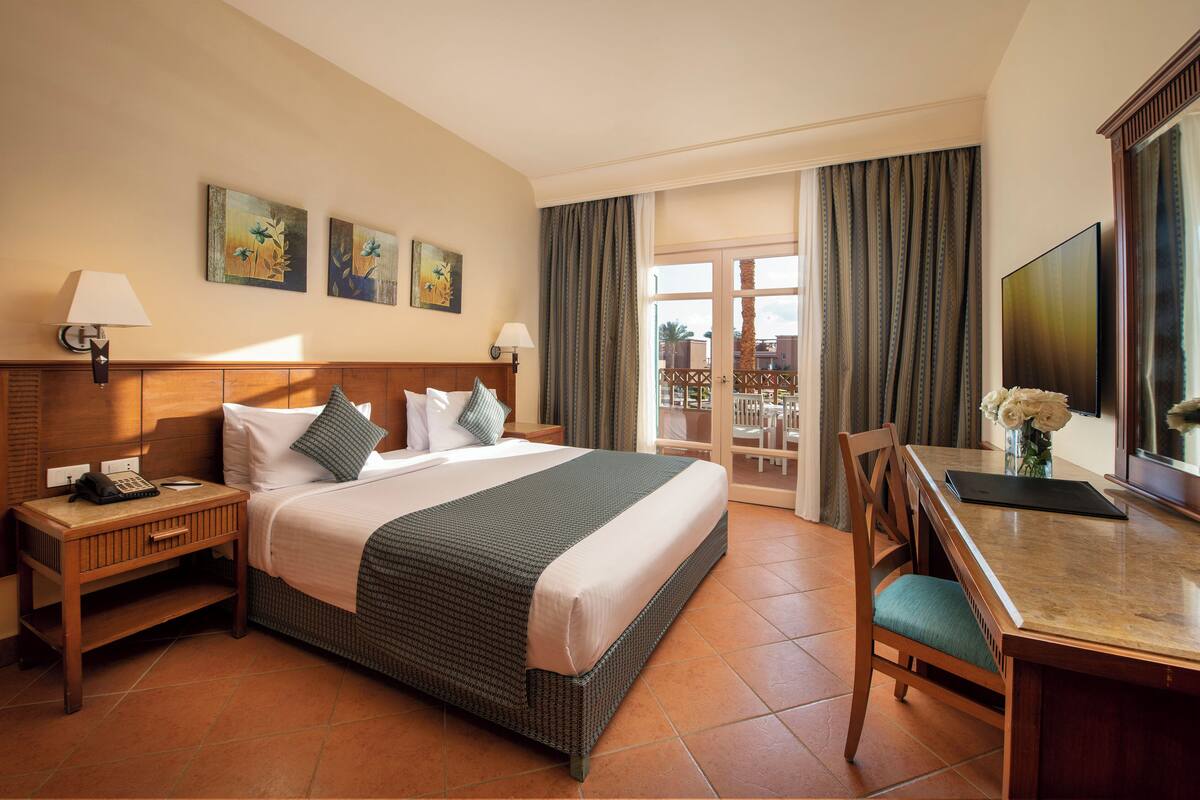 Une chambre d’hôtel comprenant un lit, des tables de chevet, un bureau avec une chaise, une télévision et un balcon avec des rideaux.