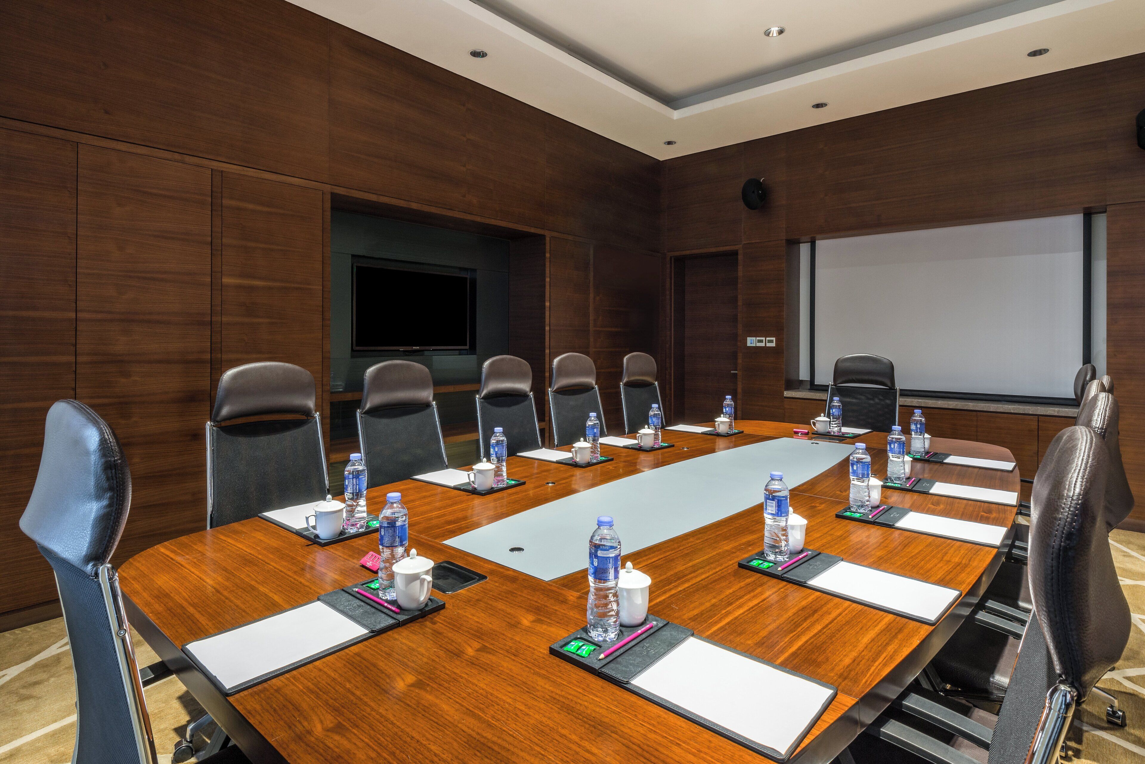 A conference room with a long wooden table, multiple chairs, water bottles, and a projector screen.