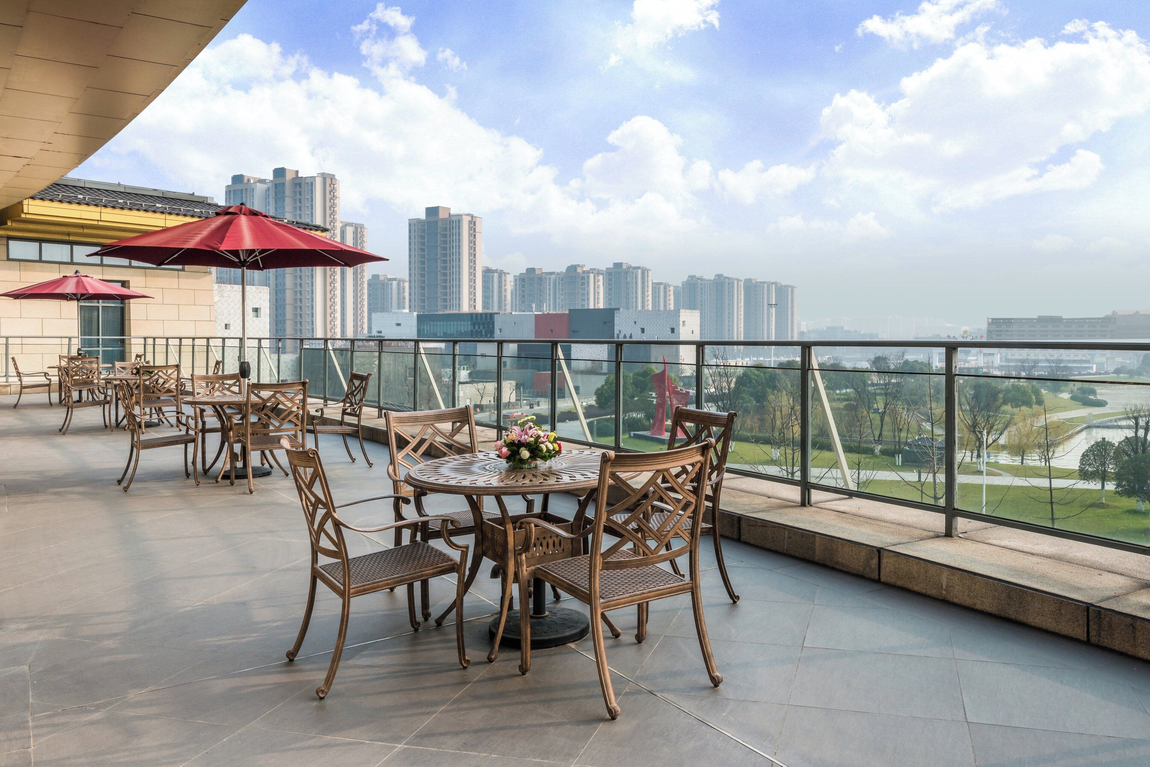Una terraza al aire libre con mesas y sillas, un paraguas rojo y vistas a un horizonte urbano.
