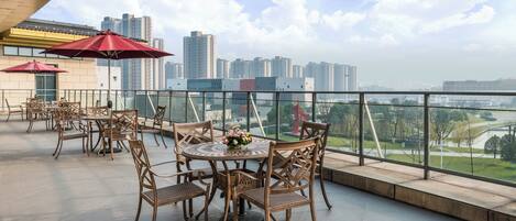 Una terraza al aire libre con mesas y sillas, un paraguas rojo y vistas a un horizonte urbano.
