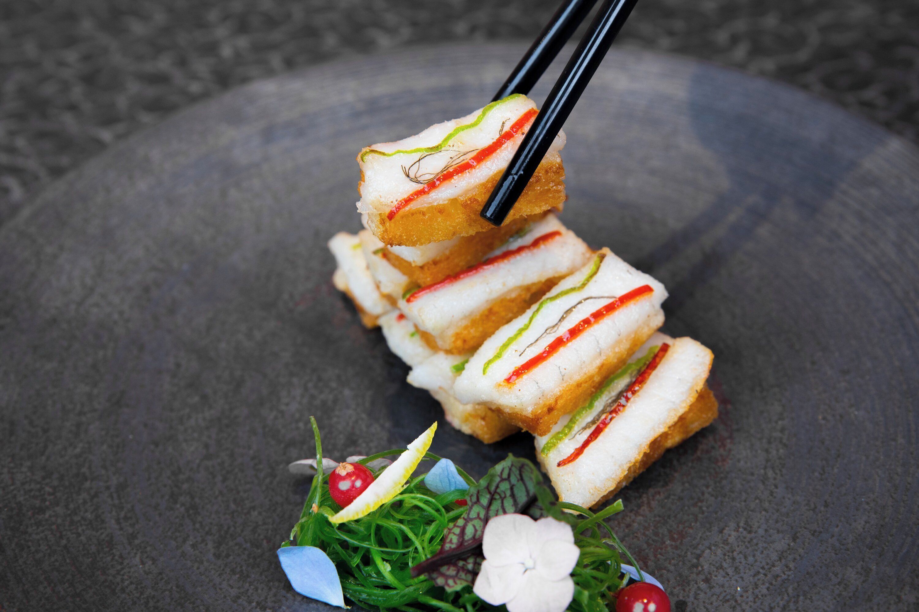 Une pile de bouchées de sushi, accompagnées de baguettes, présentées sur une assiette en pierre et décorées de feuilles vertes et d’une petite fleur.