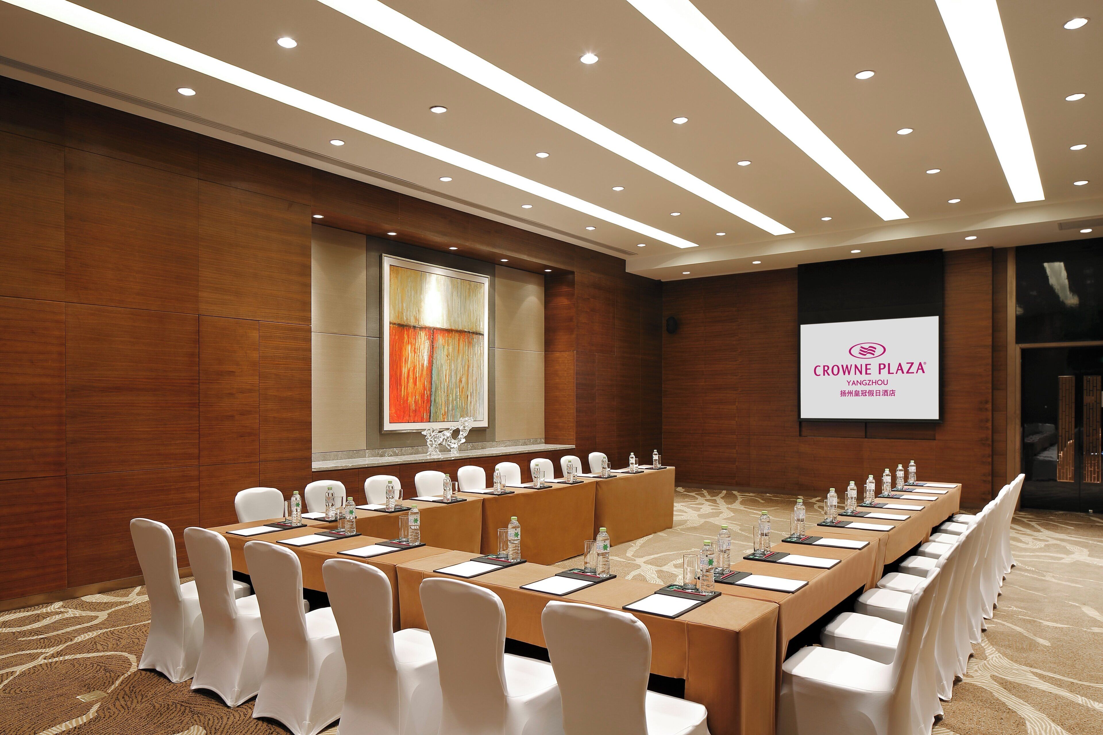 Een vergaderruimte met een lange tafel, witte stoelen en een projectorscherm waarop 'CROWNE PLAZA YANGZHOU' staat.