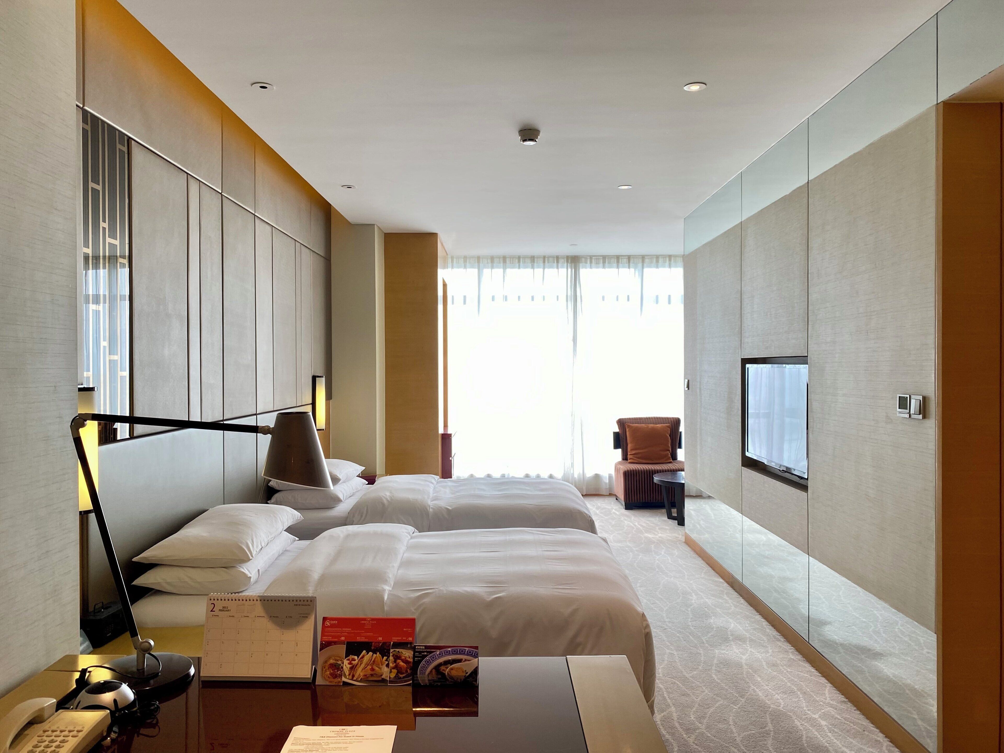 A hotel room with a bed, a desk with a lamp, a chair, a TV, and a calendar.
