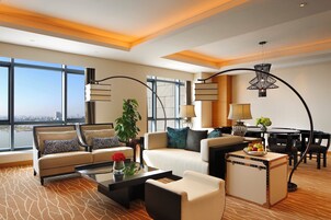 Una habitación de hotel moderna con un ventanal grande, un conjunto de sofá, una mesa de centro, una mesita auxiliar, una lámpara y un rincón comedor.