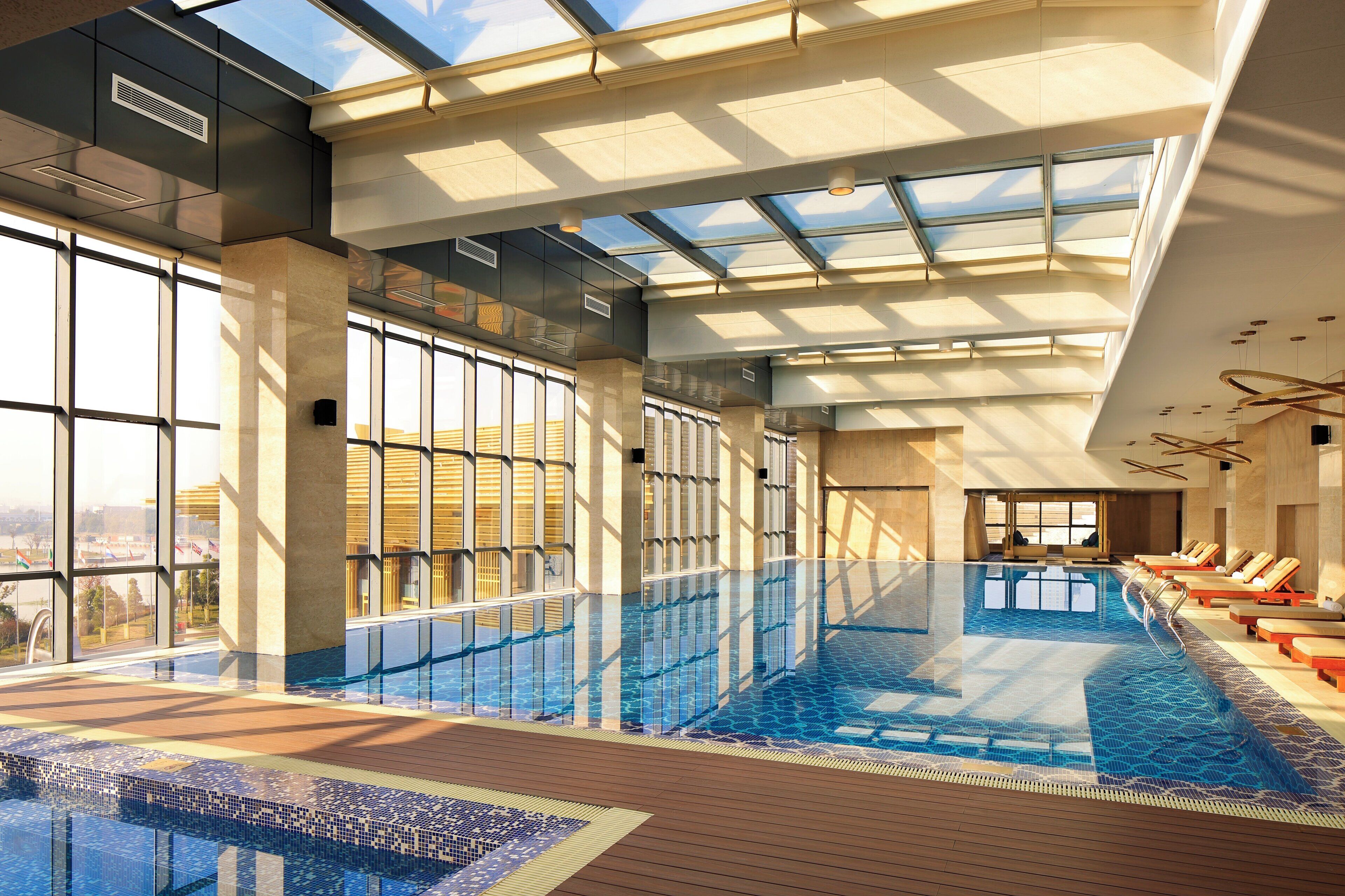 An indoor swimming pool with clear glass walls, wooden deck, and lounge chairs.