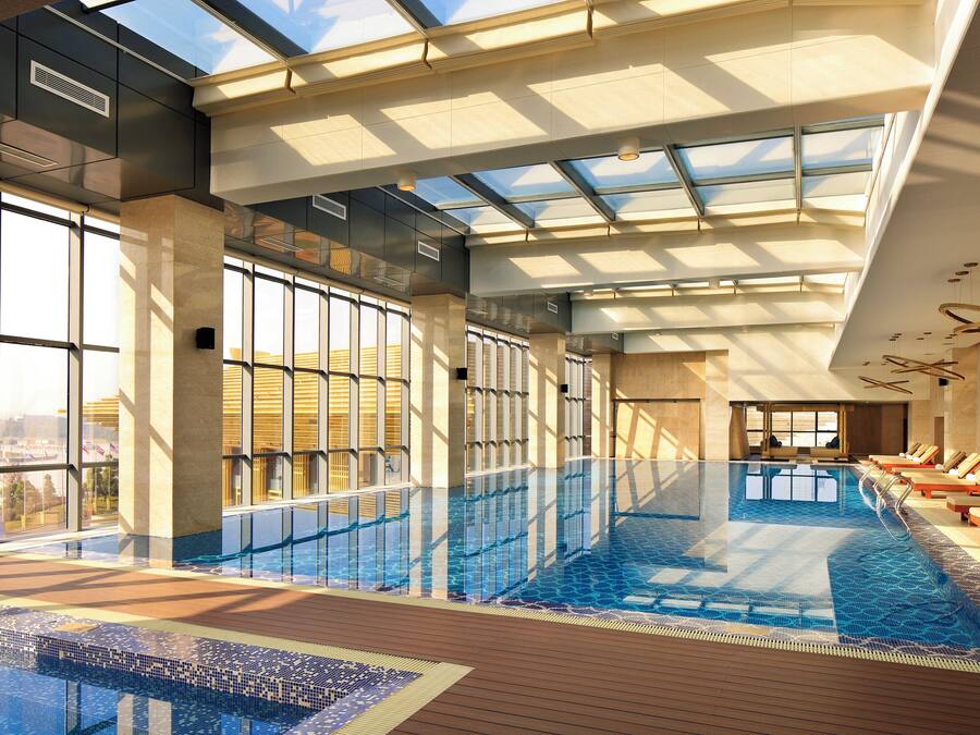 An indoor swimming pool with clear glass walls, wooden deck, and lounge chairs.