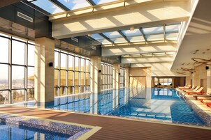 Una piscina interior con paredes de vidrio transparente, terraza de madera y sillones de descanso.