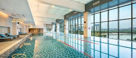 A swimming pool with a glass floor, lounge chairs, and large windows offering a view of the city.