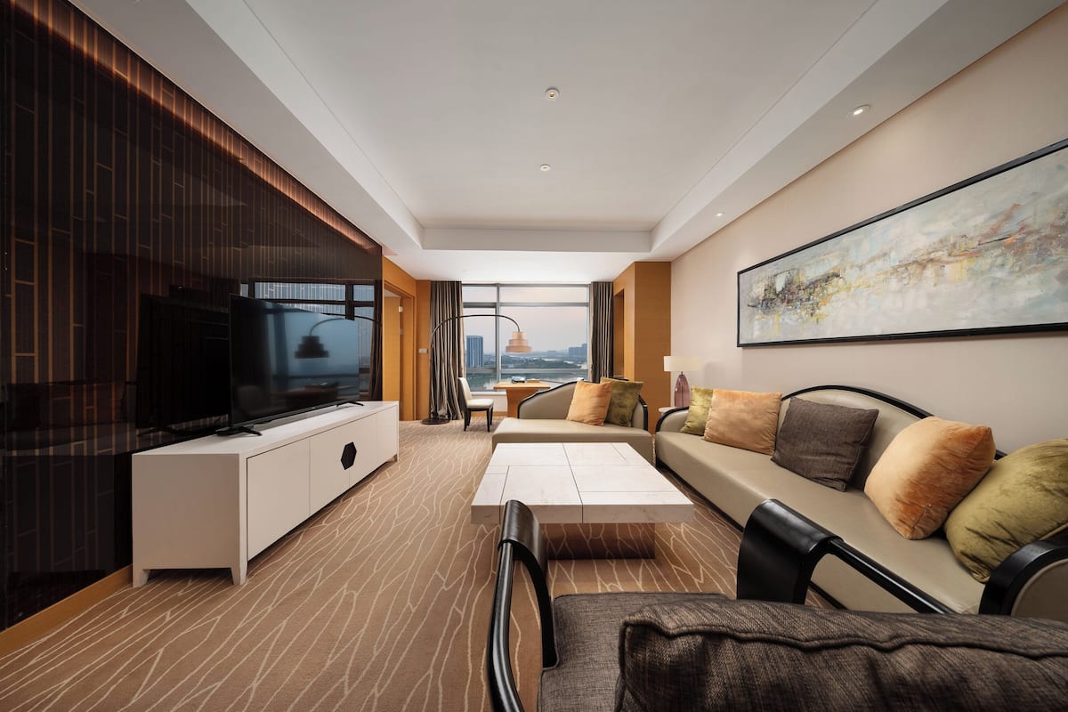 Habitación de hotel moderna con un televisor grande, un sofá con cojines, una mesa de comedor y vistas a la ciudad a través de la ventana.
