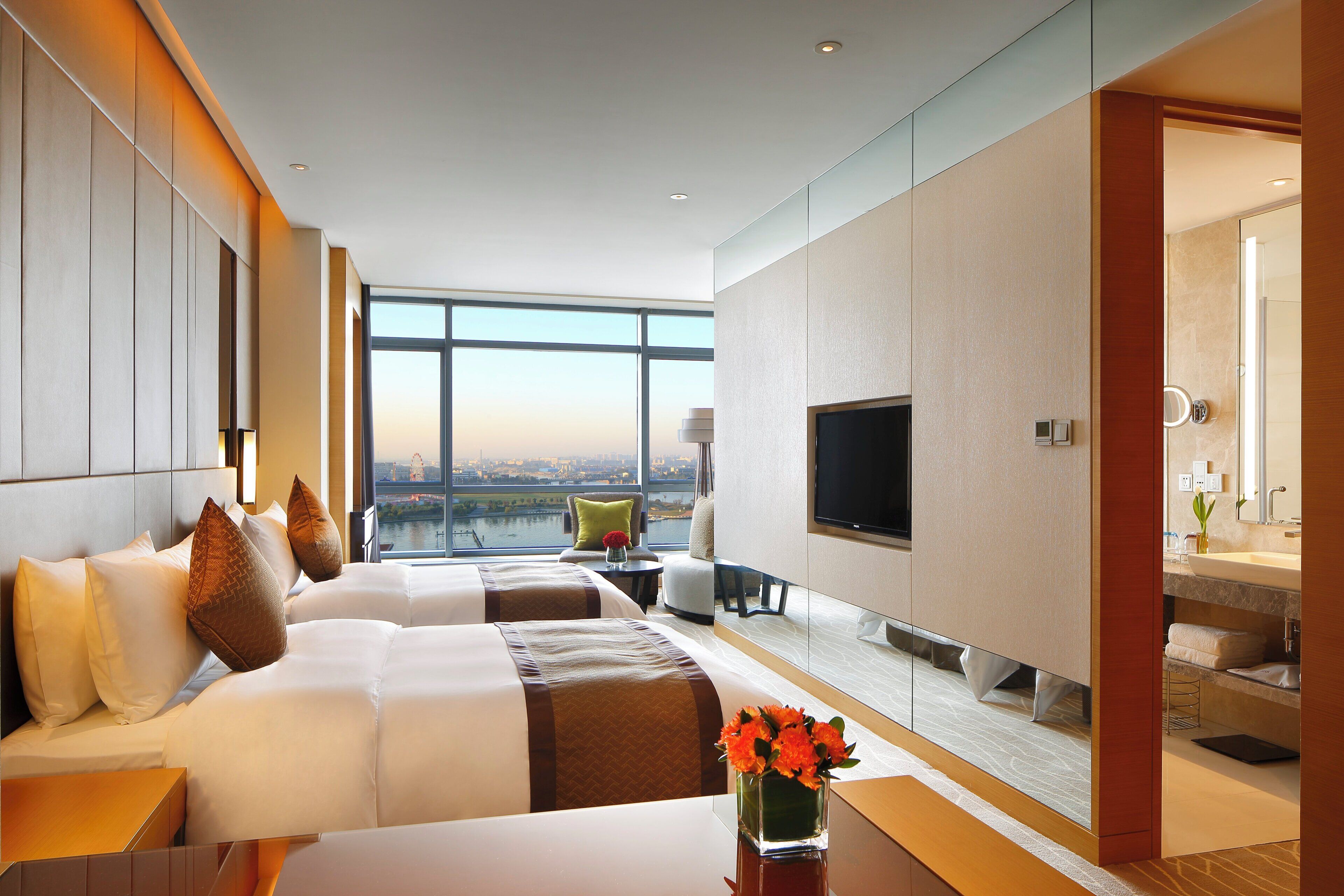 A hotel room with a large bed, a TV, a view of a cityscape, and a bathroom with a mirror and sink.