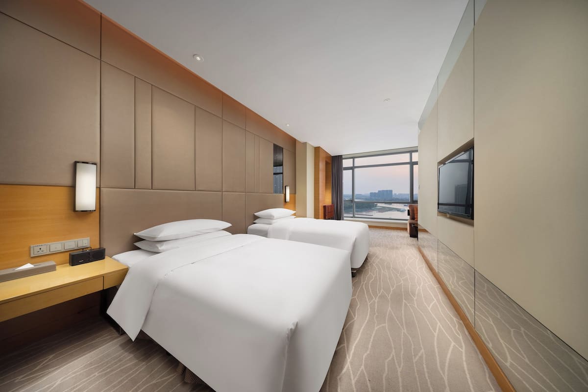 Habitación de hotel con dos camas, un escritorio y un televisor de pantalla plana, y un ventanal con vistas a la ciudad.