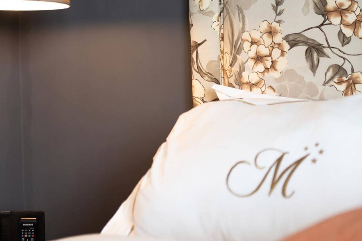 A hotel room with a dark wall, a floral wallpaper, a lamp, and a bed with a pillow featuring a monogram.