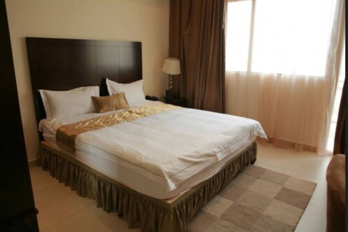 A neatly made bed with a dark headboard, a bedside lamp, and a window with curtains.
