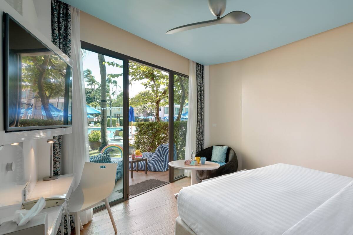 Habitación de hotel moderna con cama, escritorio y silla. Amplia ventana con vistas a una piscina y palmeras.