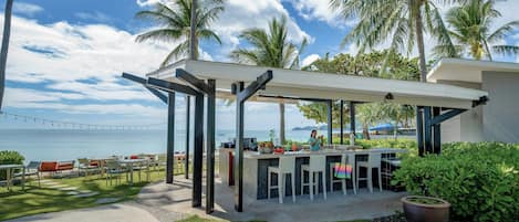 An outdoor bar area with a view of the ocean, surrounded by palm trees and a clear sky.