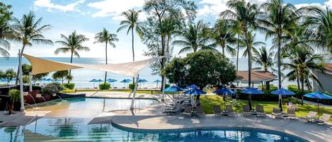 A pool area with lounge chairs and umbrellas, surrounded by palm trees and a view of the ocean.