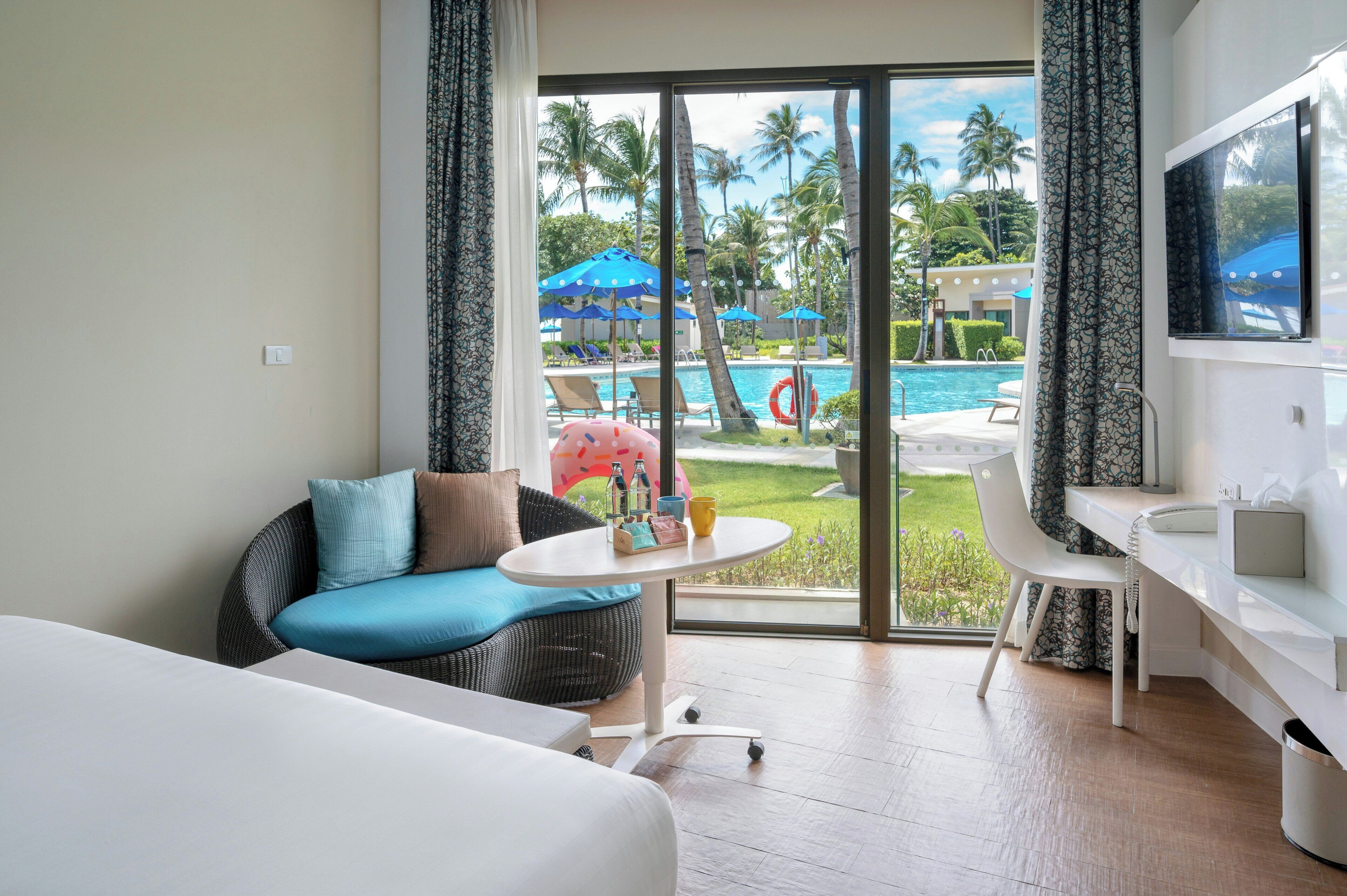 A hotel room with a bed, a sofa, a small table, a desk, a TV, and a view of a pool and palm trees.