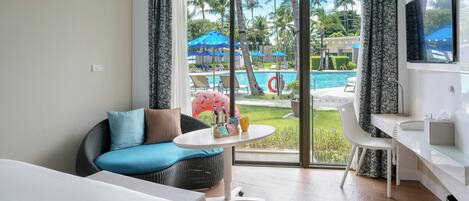 Une chambre d’hôtel avec un lit, un canapé, une petite table, un bureau, une télévision et une vue sur une piscine et des palmiers.