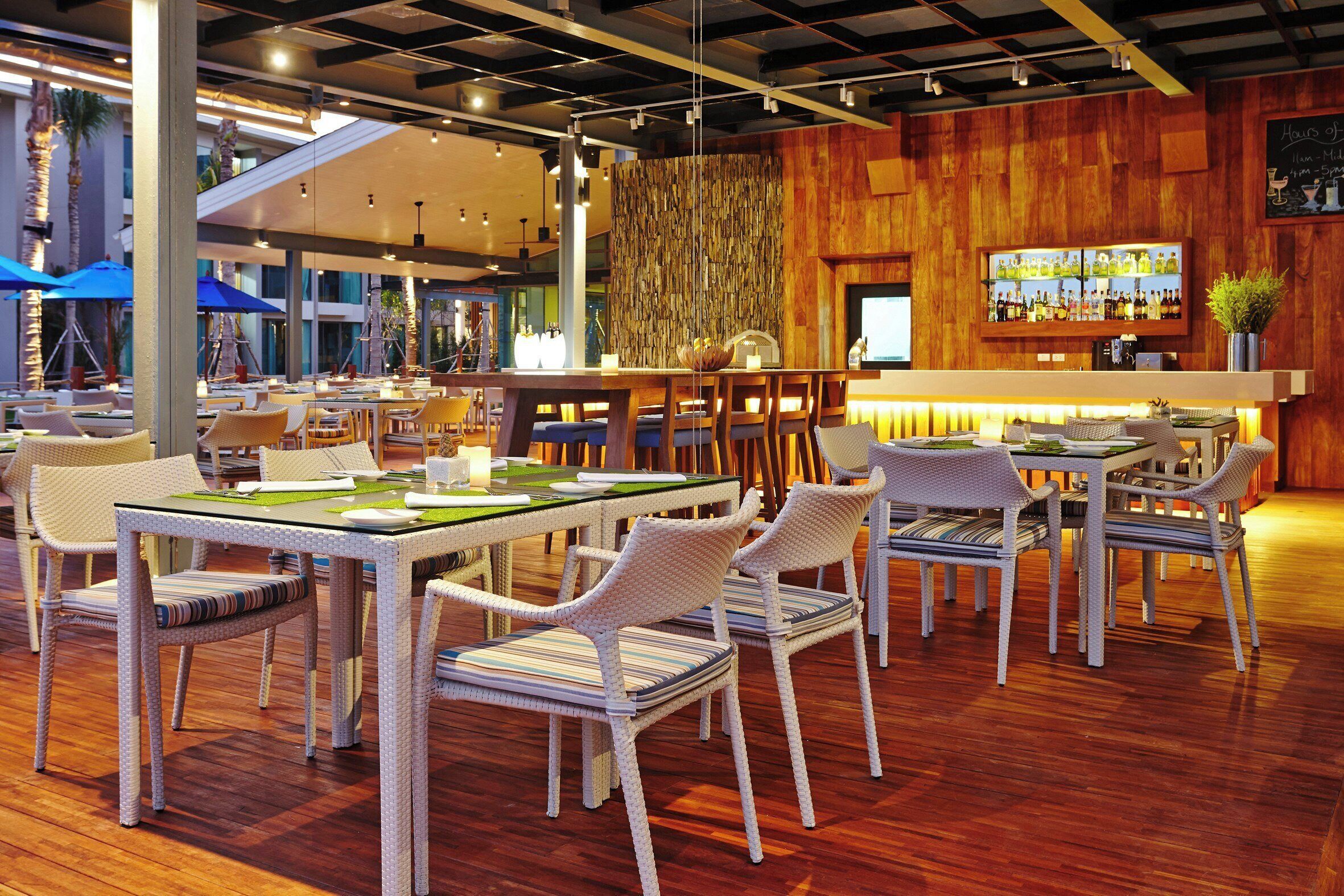 Un restaurante con sillas de mimbre, mesas de madera, zona de bar y pared de madera.