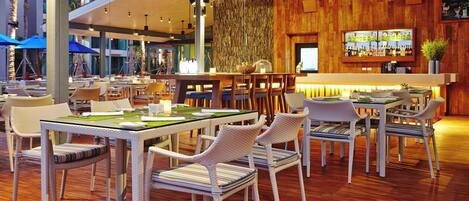 Un restaurante con sillas de mimbre, mesas de madera, zona de bar y pared de madera.
