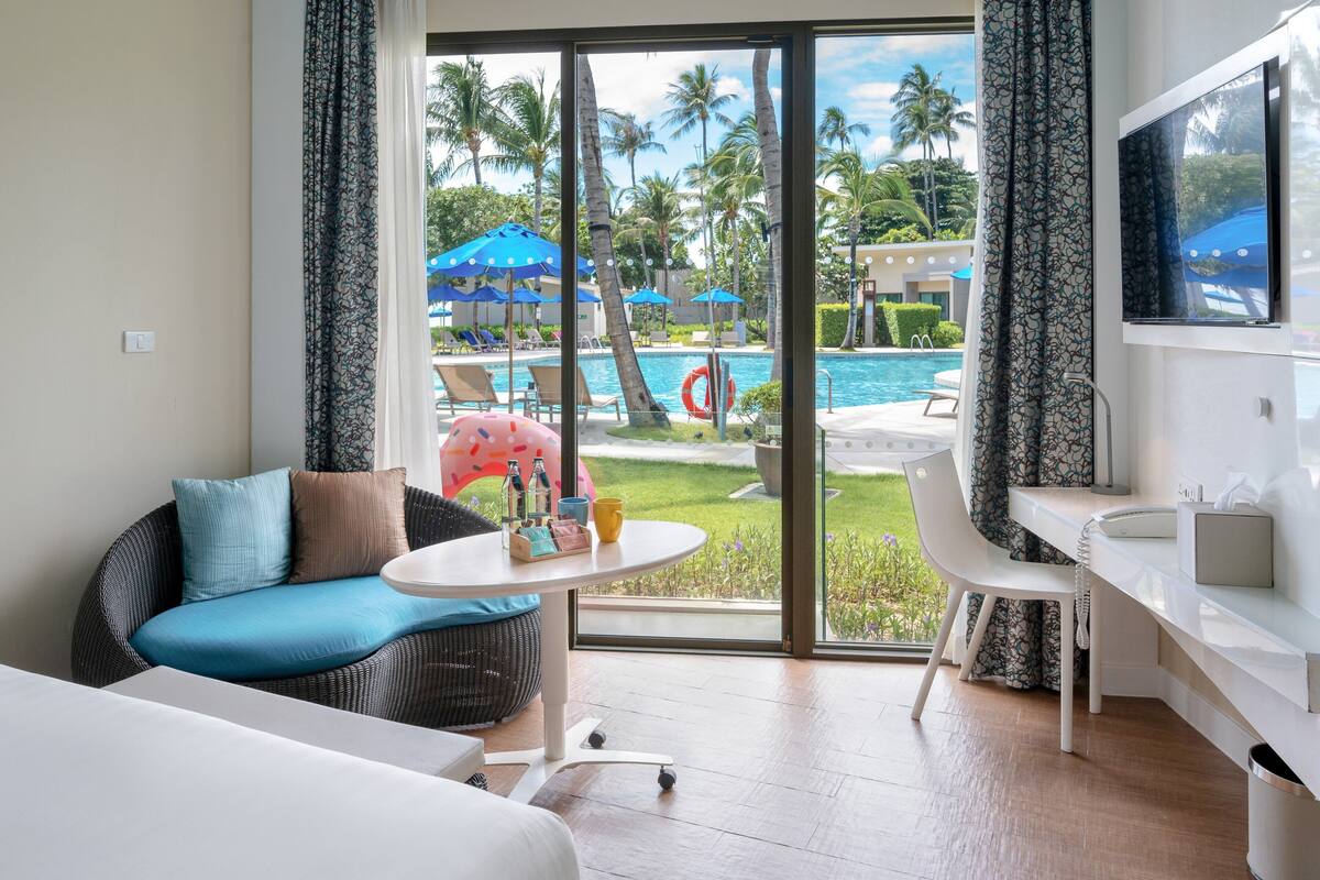 Habitación de hotel con sofá, una mesita redonda, televisor y vista a un área de piscina con sombrillas y palmeras.