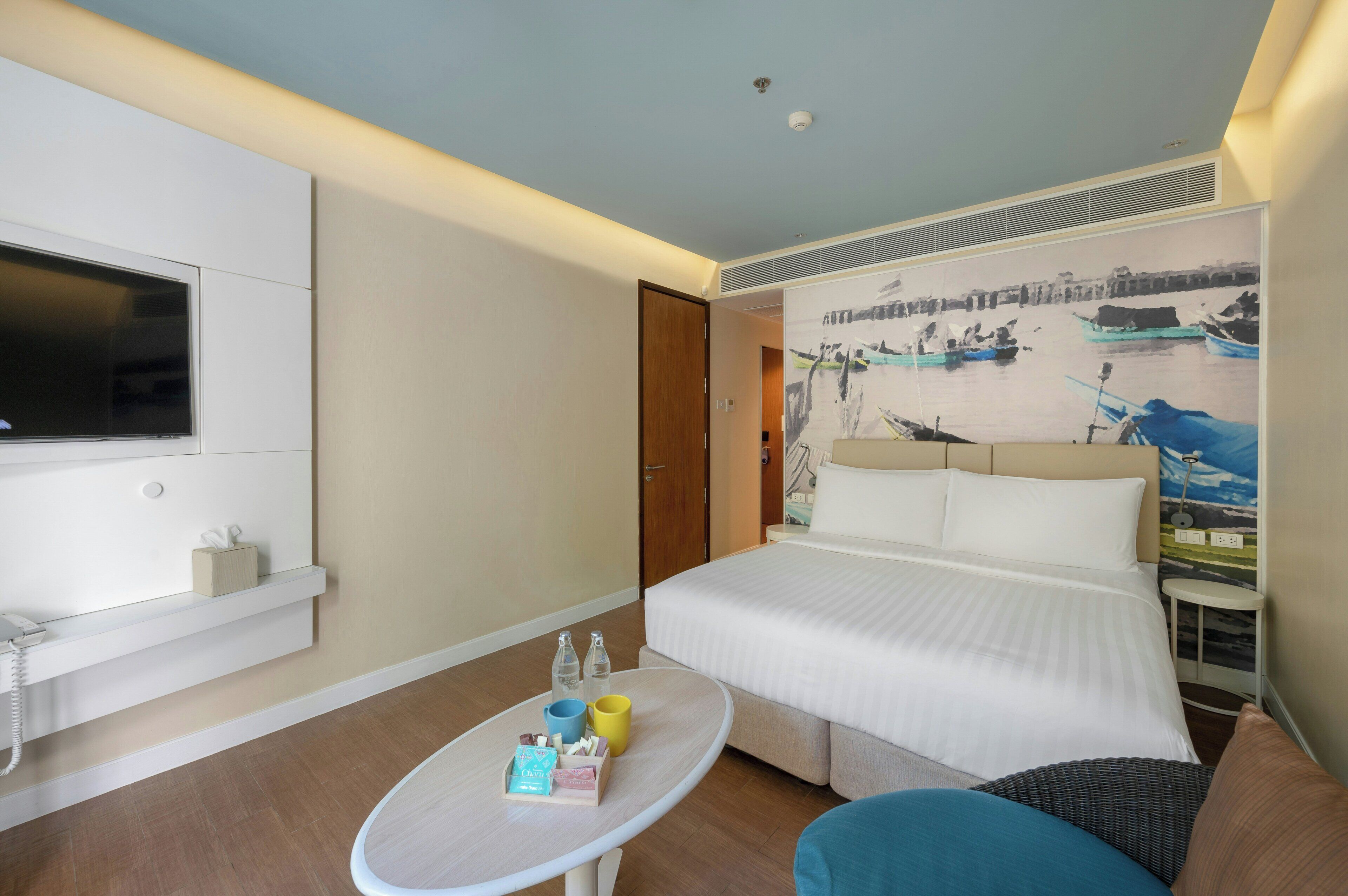 Habitación de hotel con cama, televisión montada en la pared, una mesita con artículos de aseo y un cuadro de barcos en la pared.