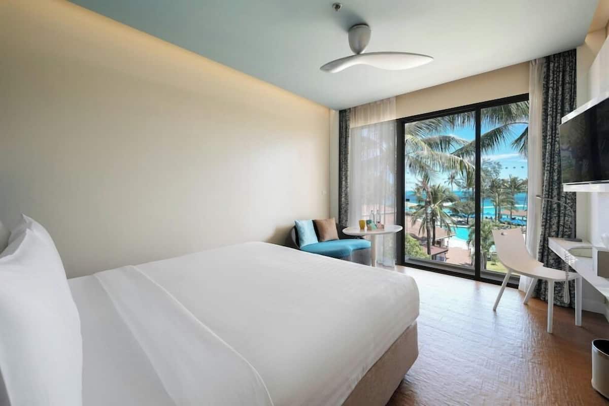 Habitación de hotel con cama, escritorio, silla y ventana grande con vistas a una piscina y palmeras.