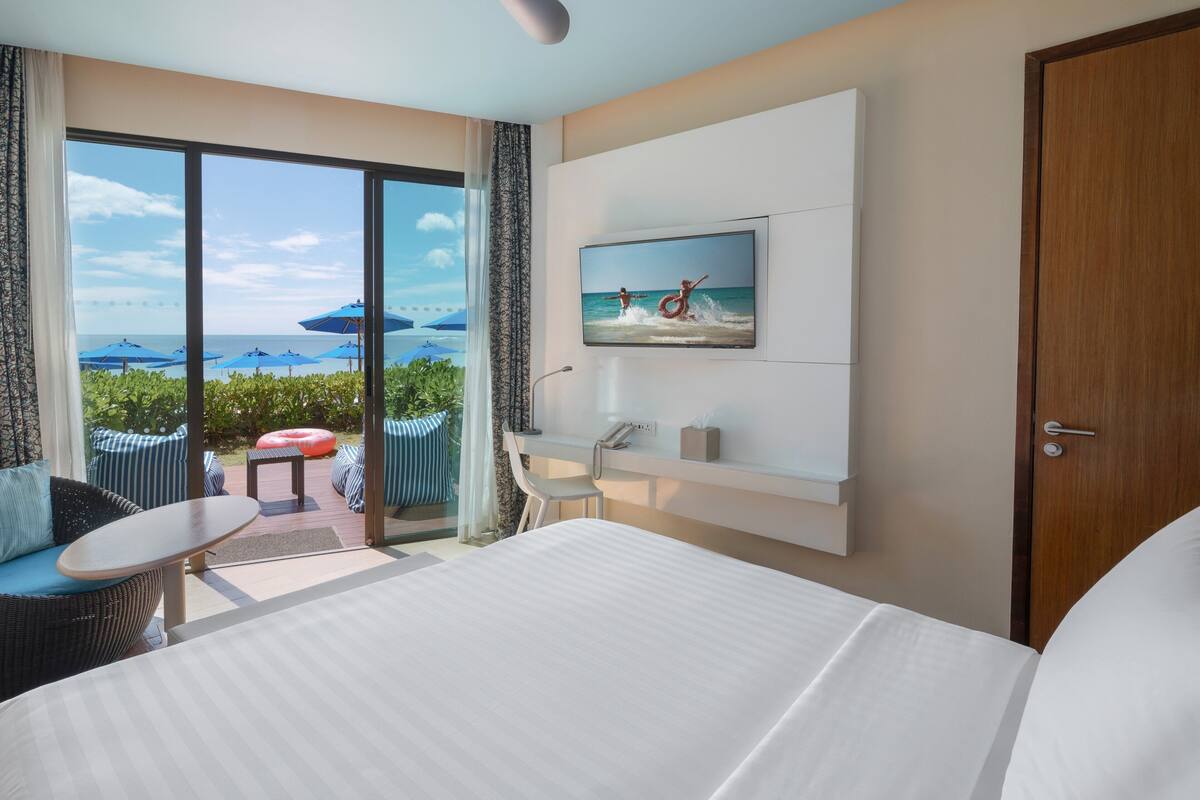 Habitación de hotel con una cama grande, televisor montado en la pared, balcón con mobiliario exterior y vista al mar.