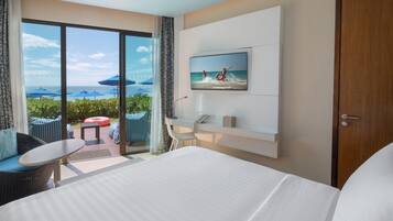 A hotel room with a large bed, a TV mounted on the wall, a balcony with outdoor seating, and a view of the sea.