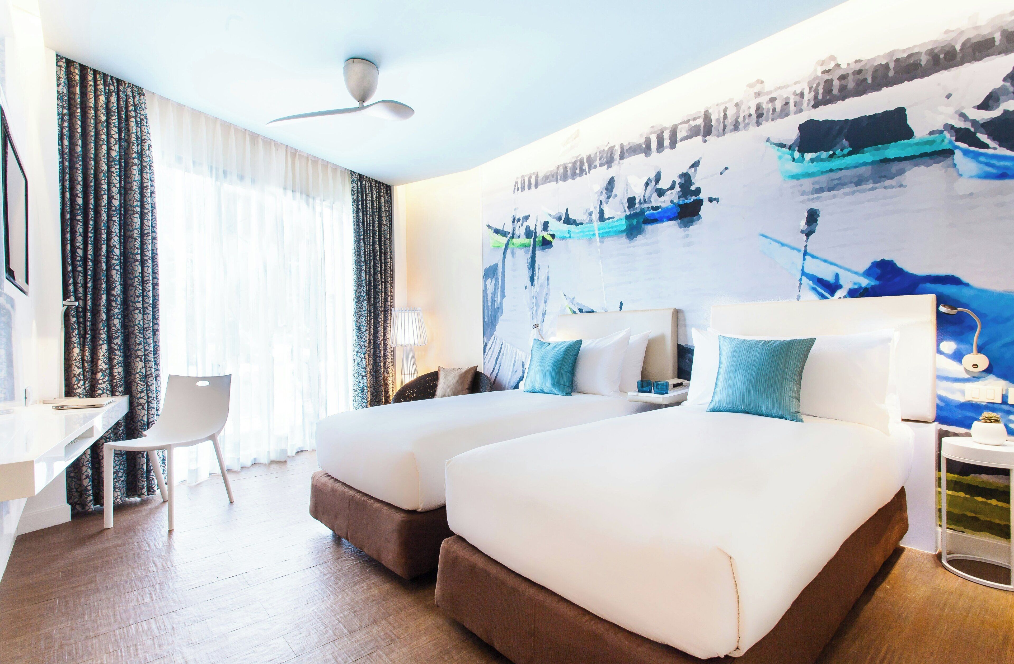 A hotel room with two beds, a desk, a chair, a ceiling fan, and a large abstract painting on the wall.