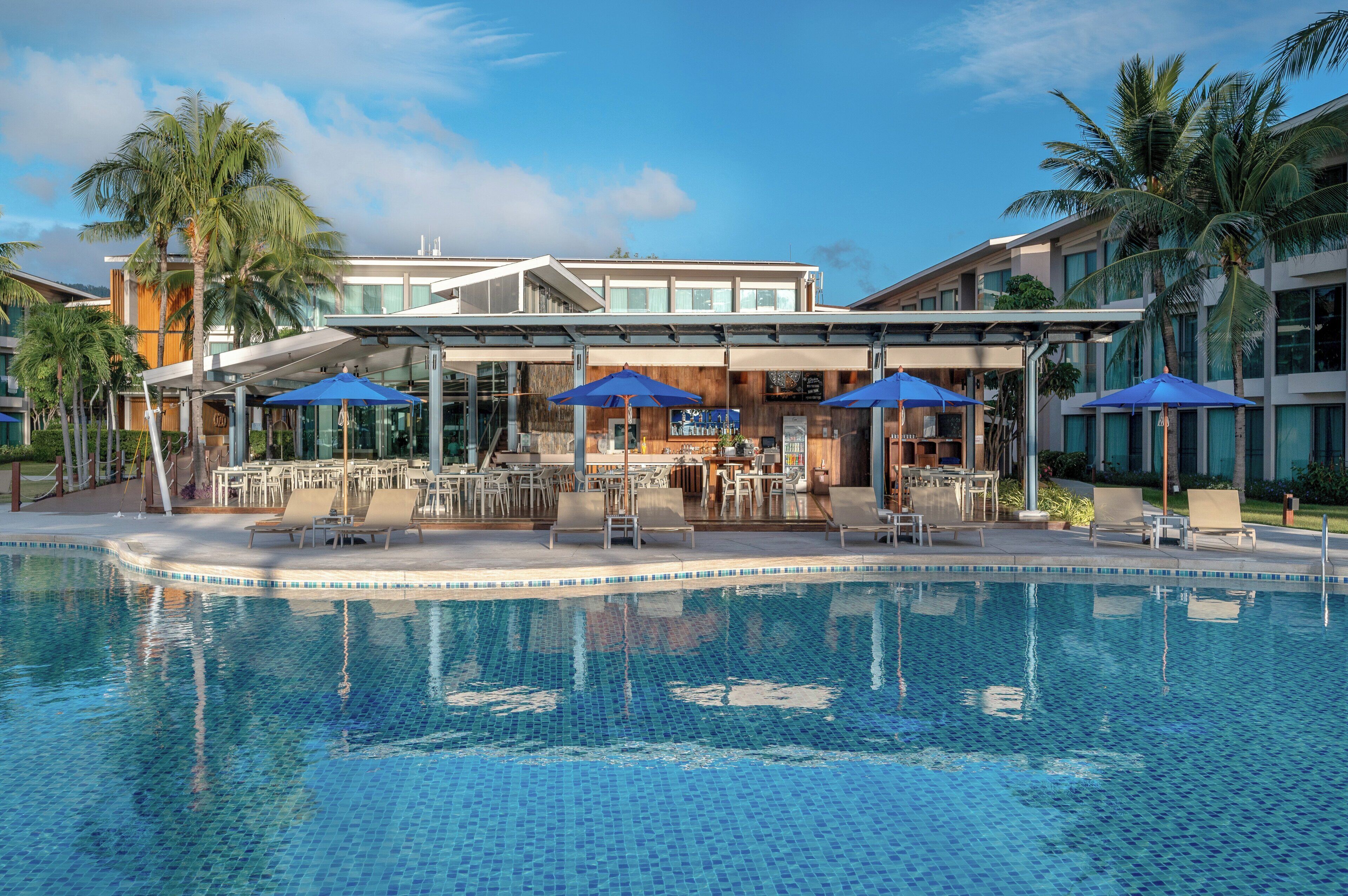 A swimming pool with lounge chairs and umbrellas, surrounded by a modern building with large windows and palm trees.