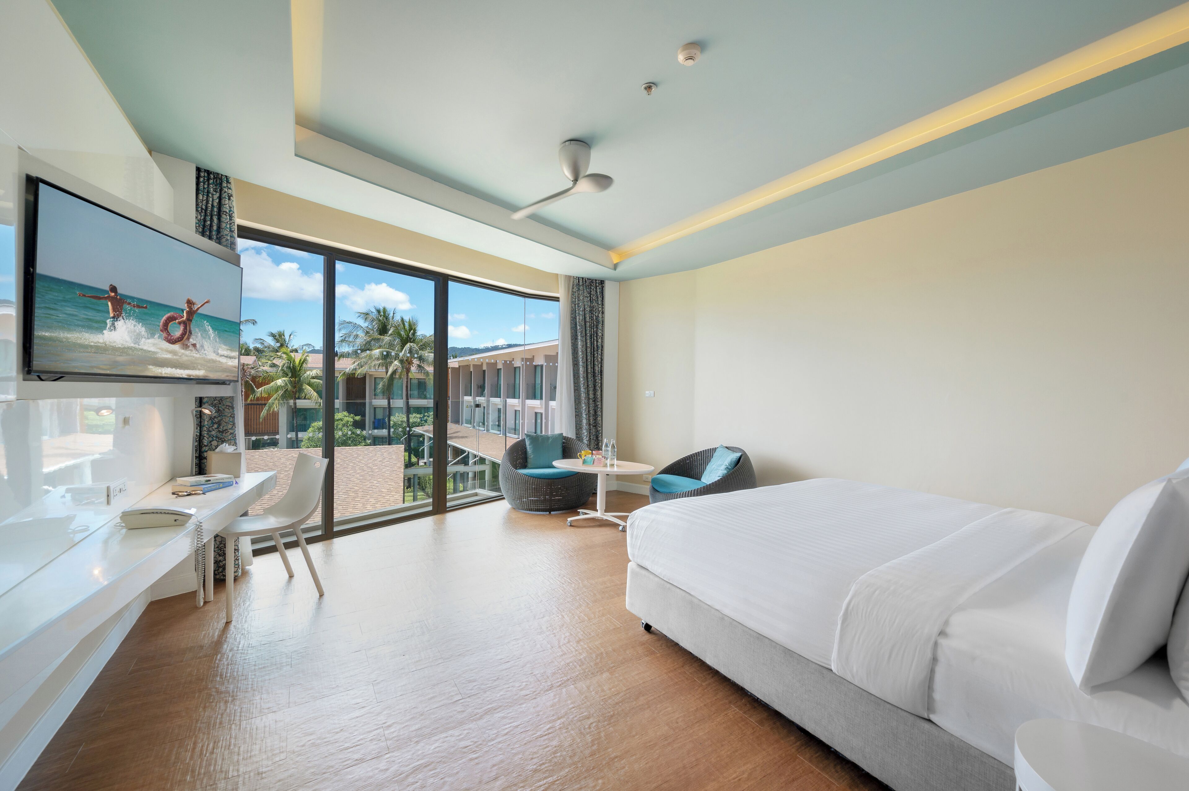 Une chambre d’hôtel moderne dotée d’un grand lit, d’un bureau, d’une chaise, d’une télévision et d’un balcon offrant une vue sur des palmiers et un immeuble.