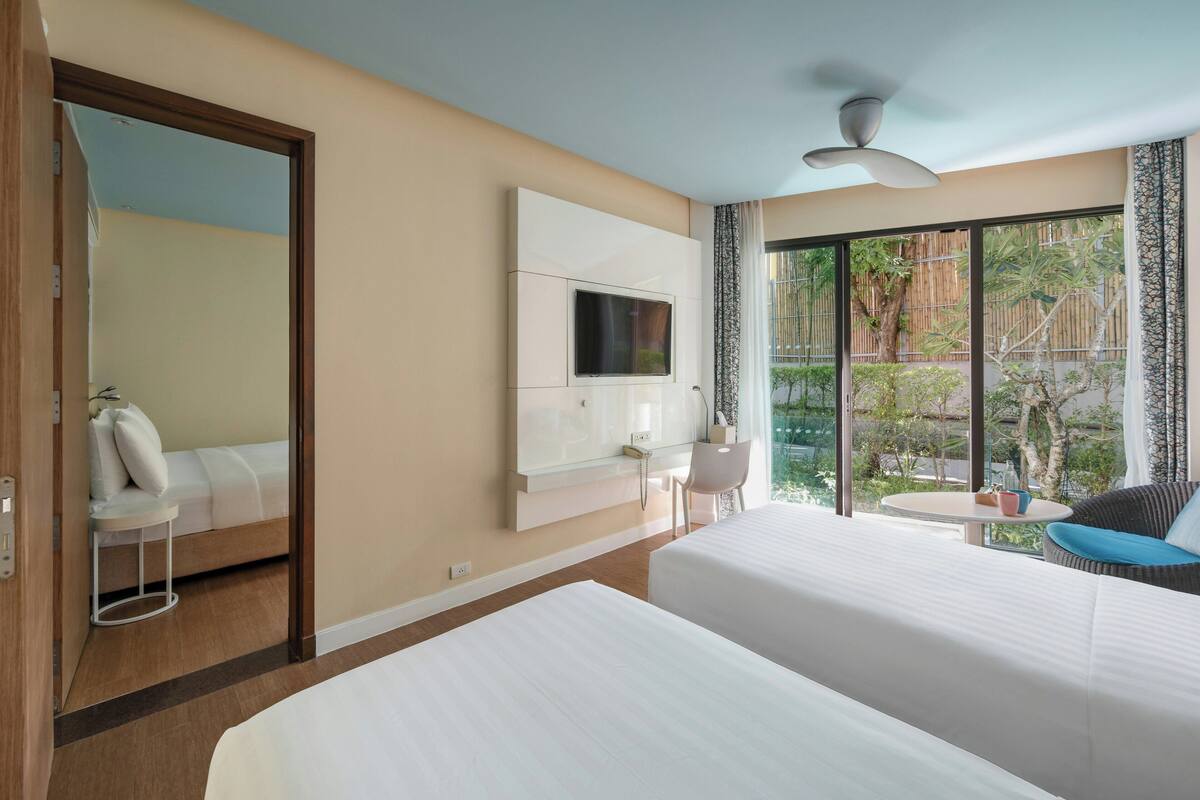 Habitación de hotel con dos camas, televisión, ventilador de techo, ventana con vistas a follaje y una mesita con una silla.