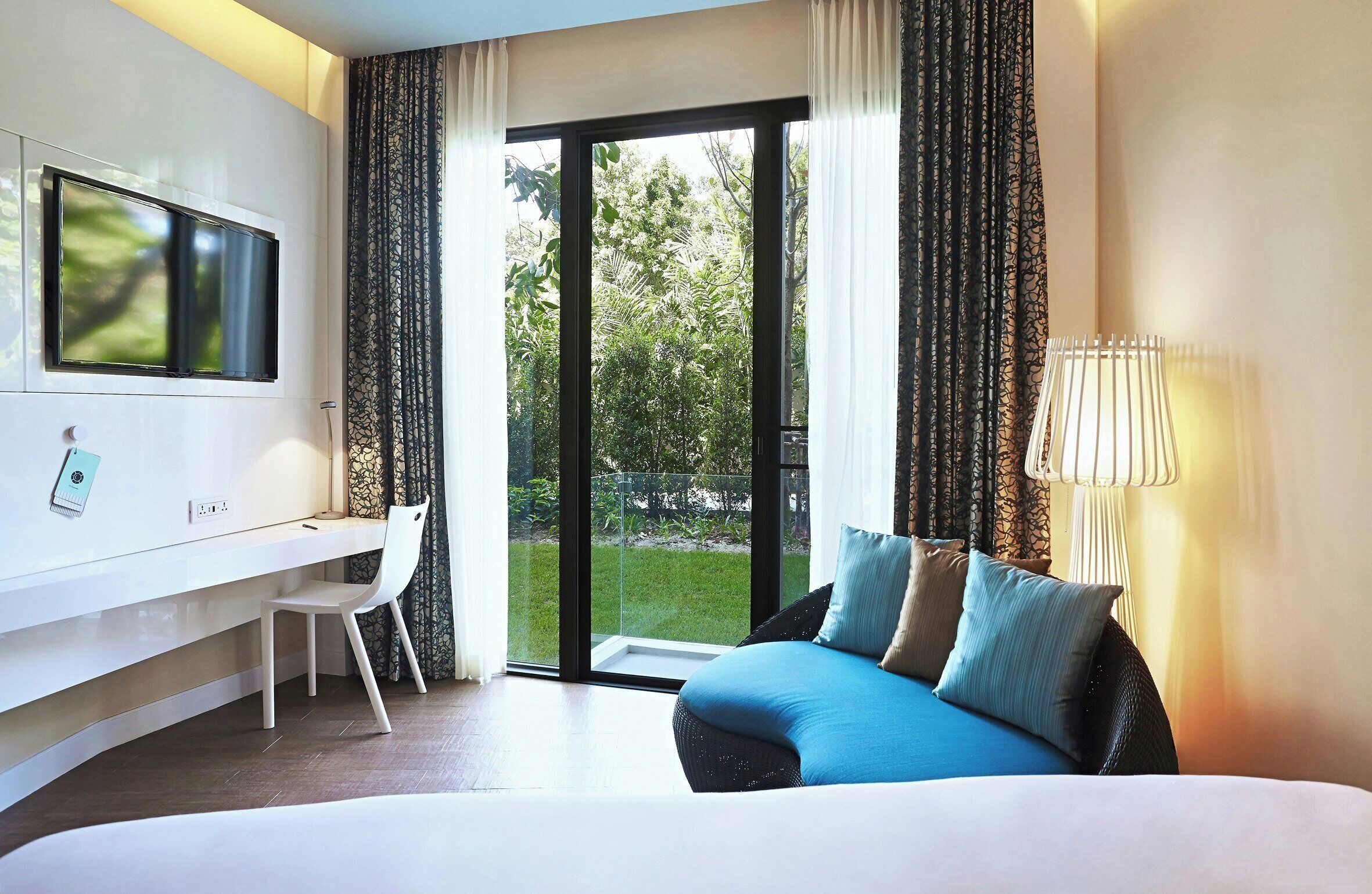 A hotel room with a bed, a desk, a chair, a TV, and a view of greenery through the glass door.