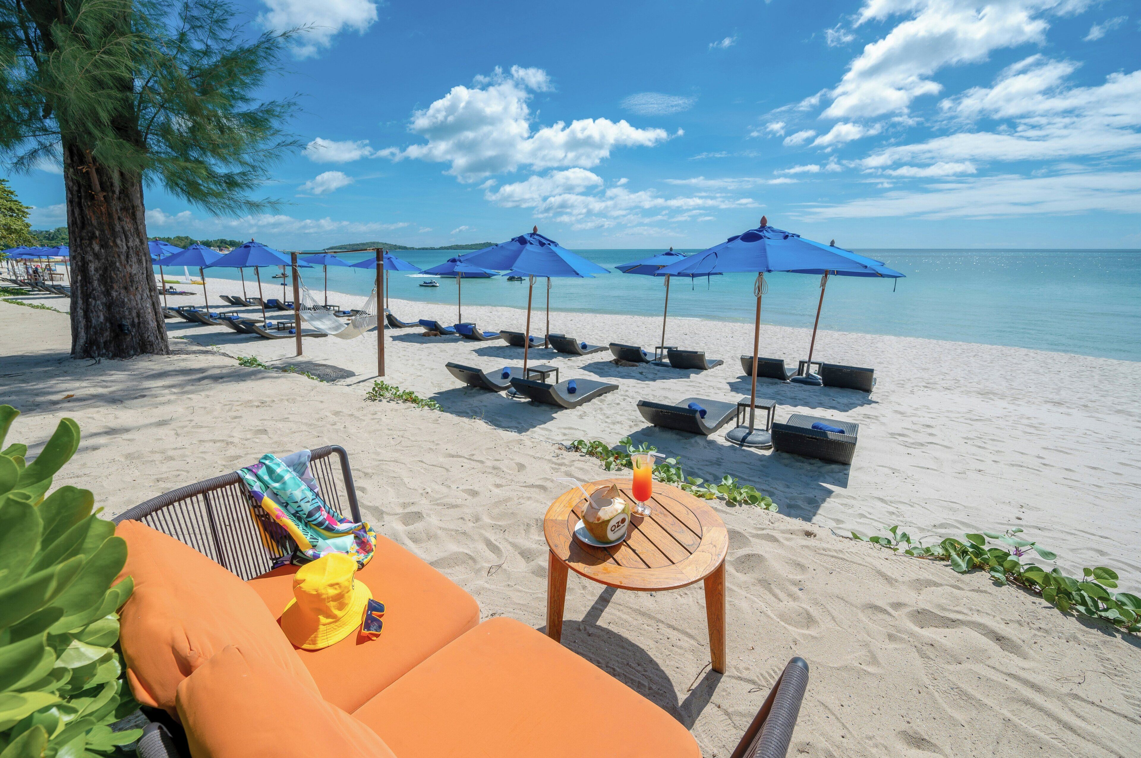 A beach with lounge chairs, umbrellas, and a table with a drink and a hat.