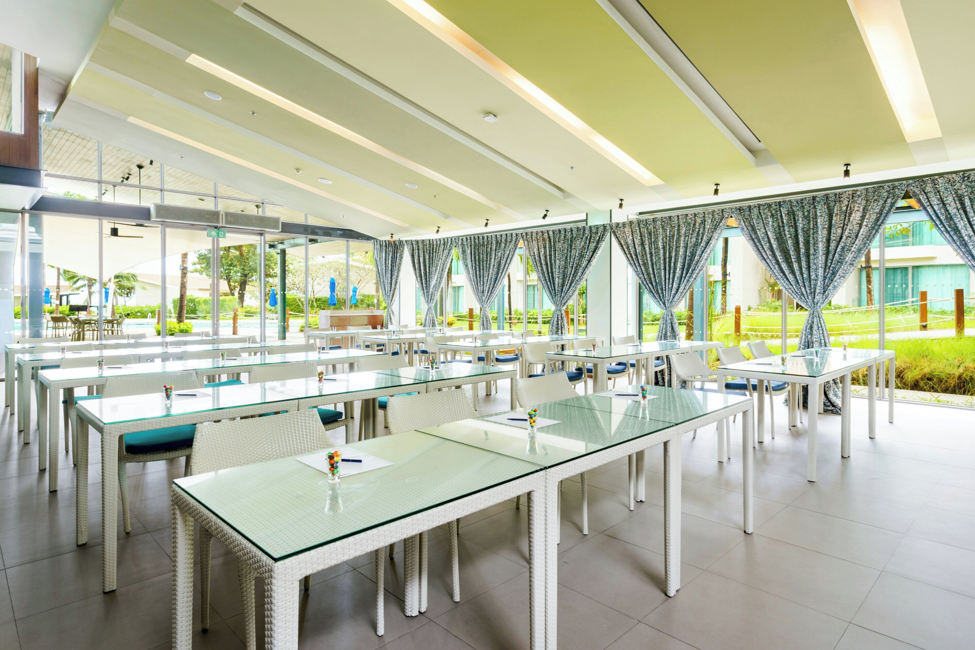 A spacious indoor dining area with glass tables and white chairs, large windows with curtains, and a view of greenery outside.