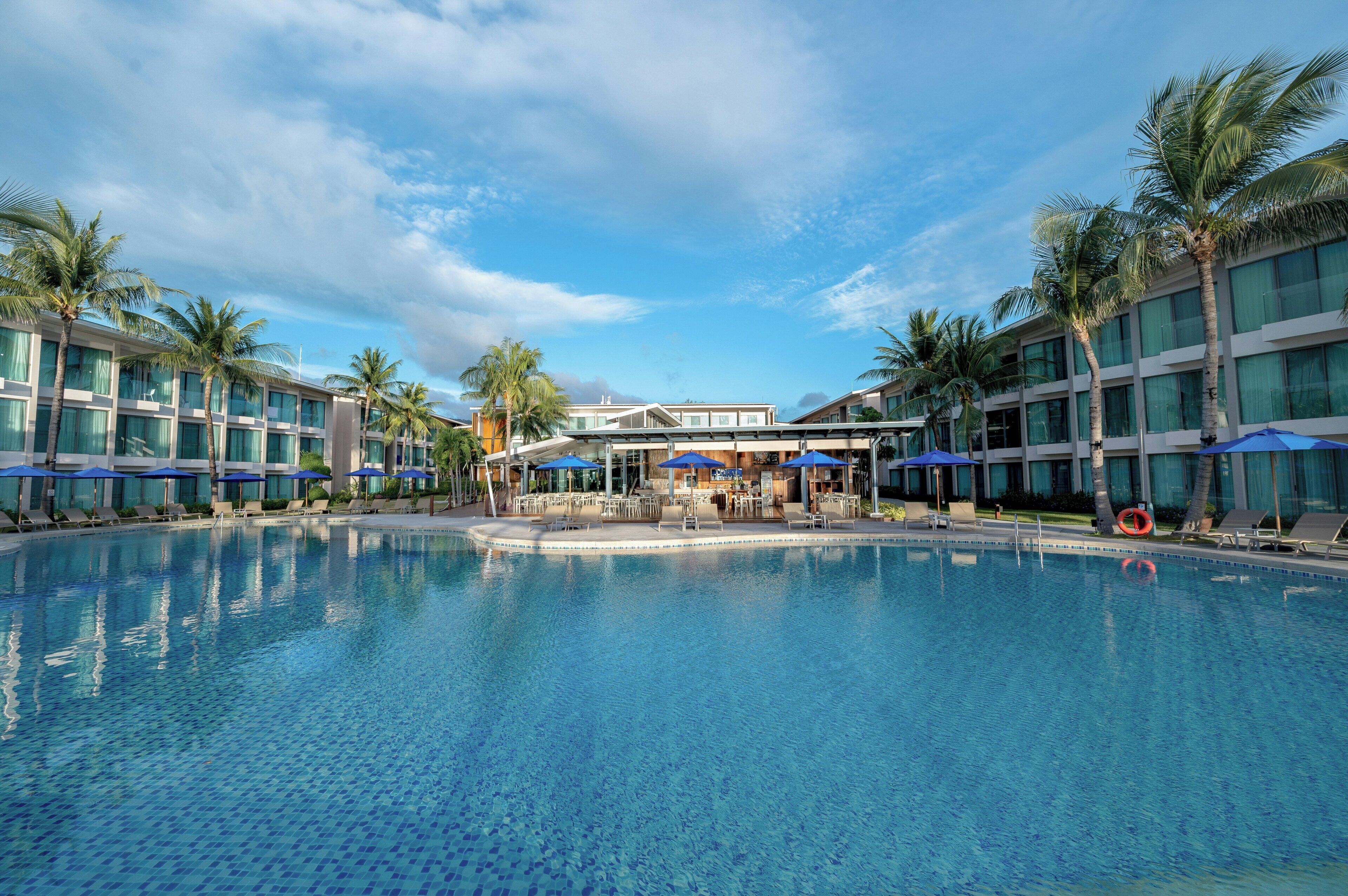 A swimming pool with lounge chairs and umbrellas, surrounded by palm trees and modern buildings.