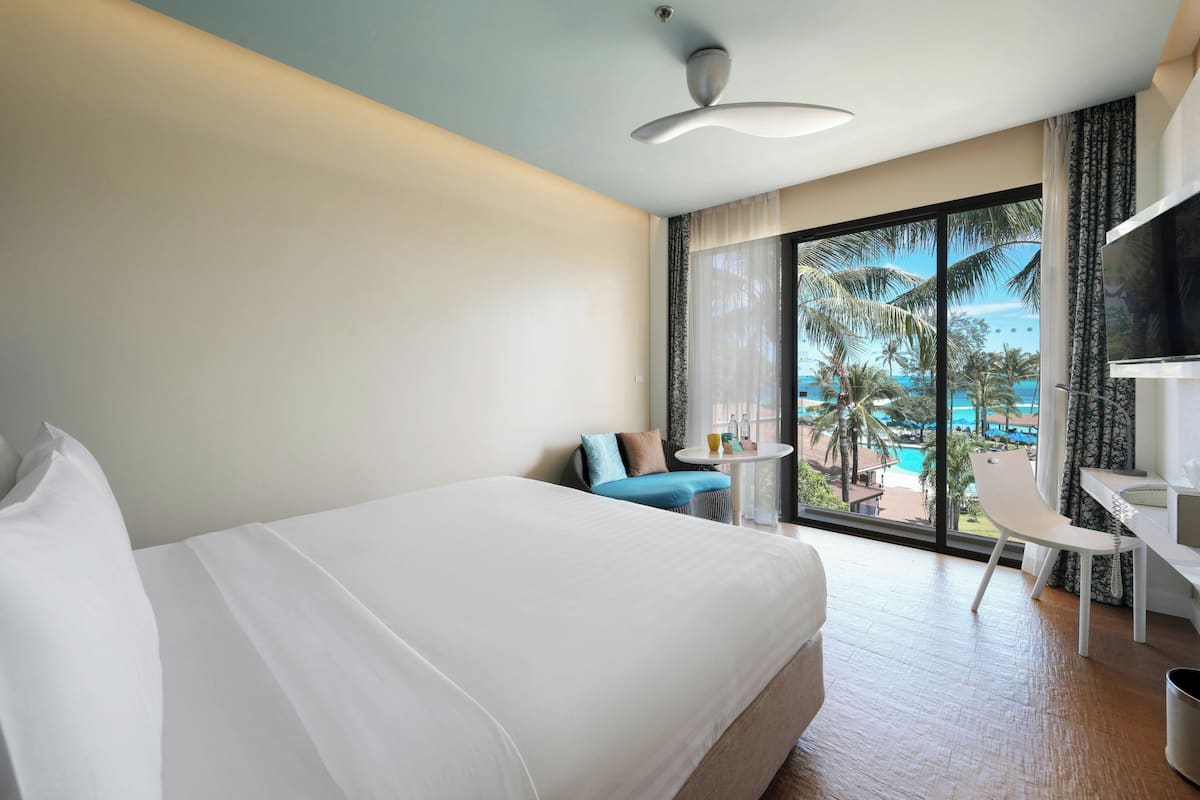 Habitación de hotel con cama, escritorio, silla, mesita pequeña y ventana grande con vistas a una piscina y palmeras.