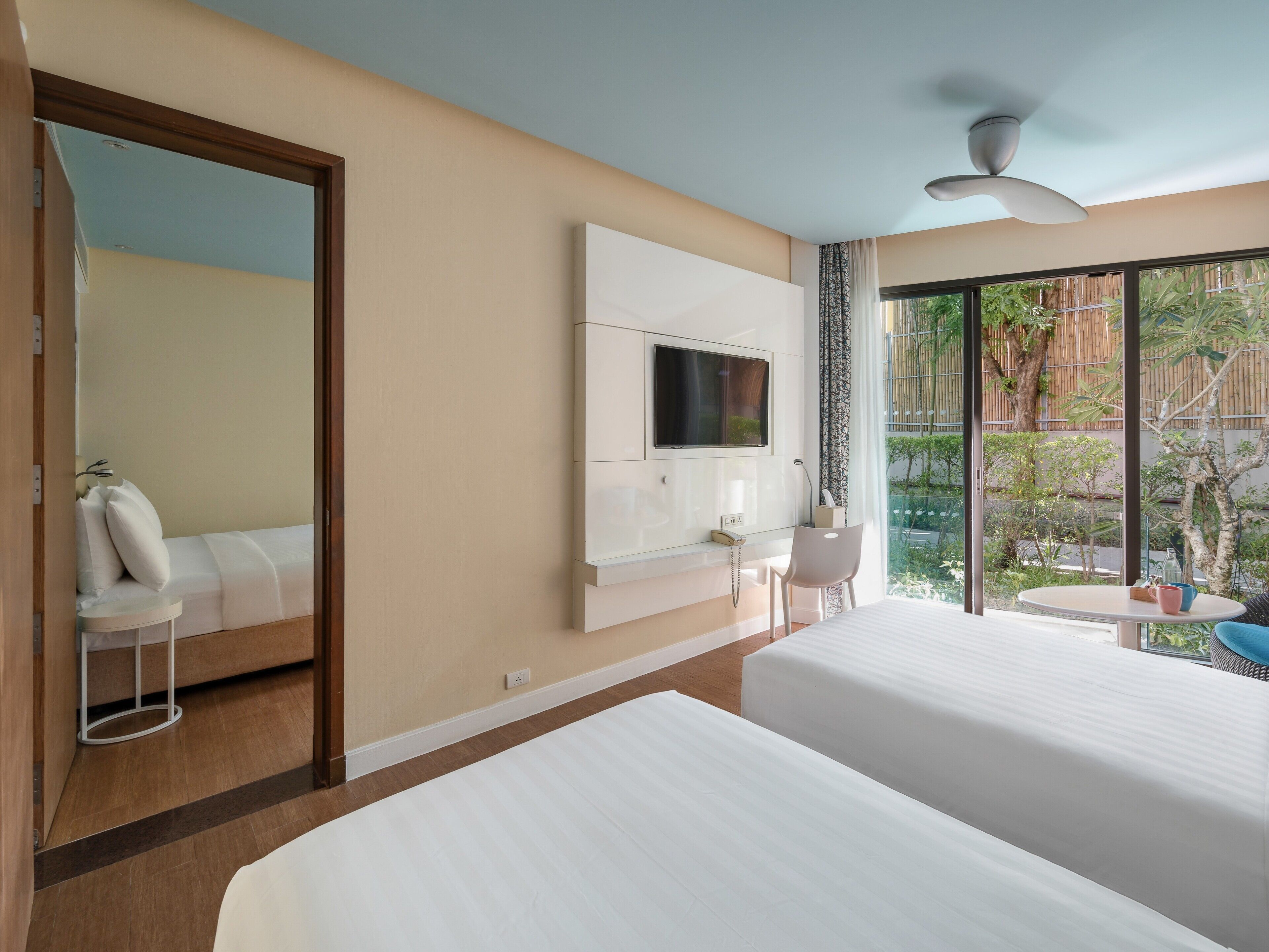 Habitación de hotel con dos camas, un televisor montado en la pared, un escritorio con silla y un ventanal con vistas a zonas verdes.